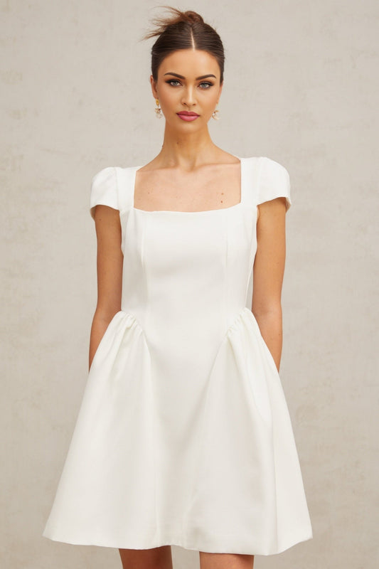 A white mini dress with cap sleeves worn by a model standing in a neutral background The dress features a fitted bodice and a flared skirt with a short hemline