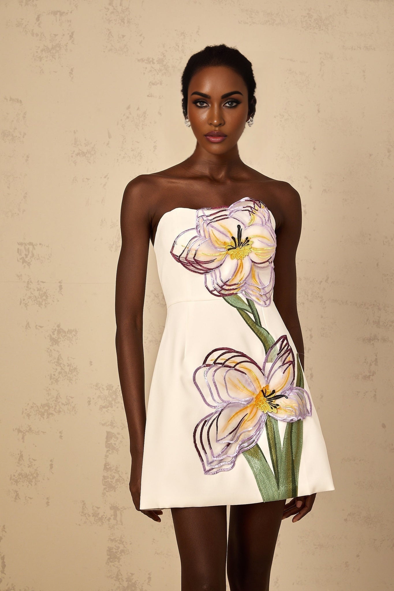 A white mini dress with offshoulder design featuring sequin floral embroidery worn by a woman standing in a neutral setting