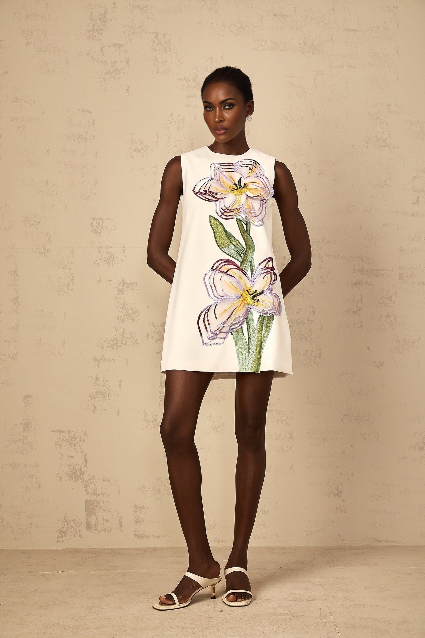 A white mini dress with sequin floral embroidery worn by a woman standing in a neutral background