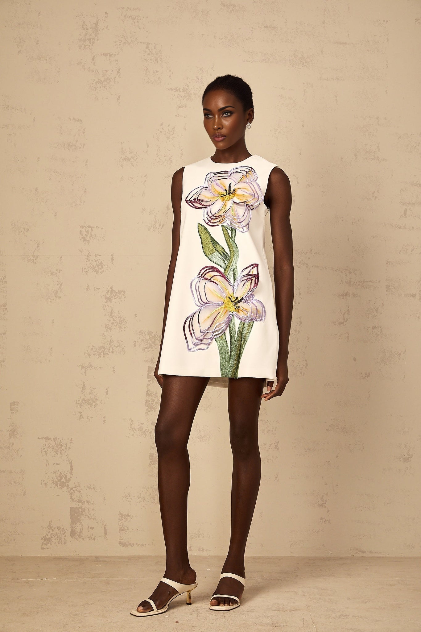 A white mini dress with sequin floral embroidery worn by a woman standing in a neutral setting