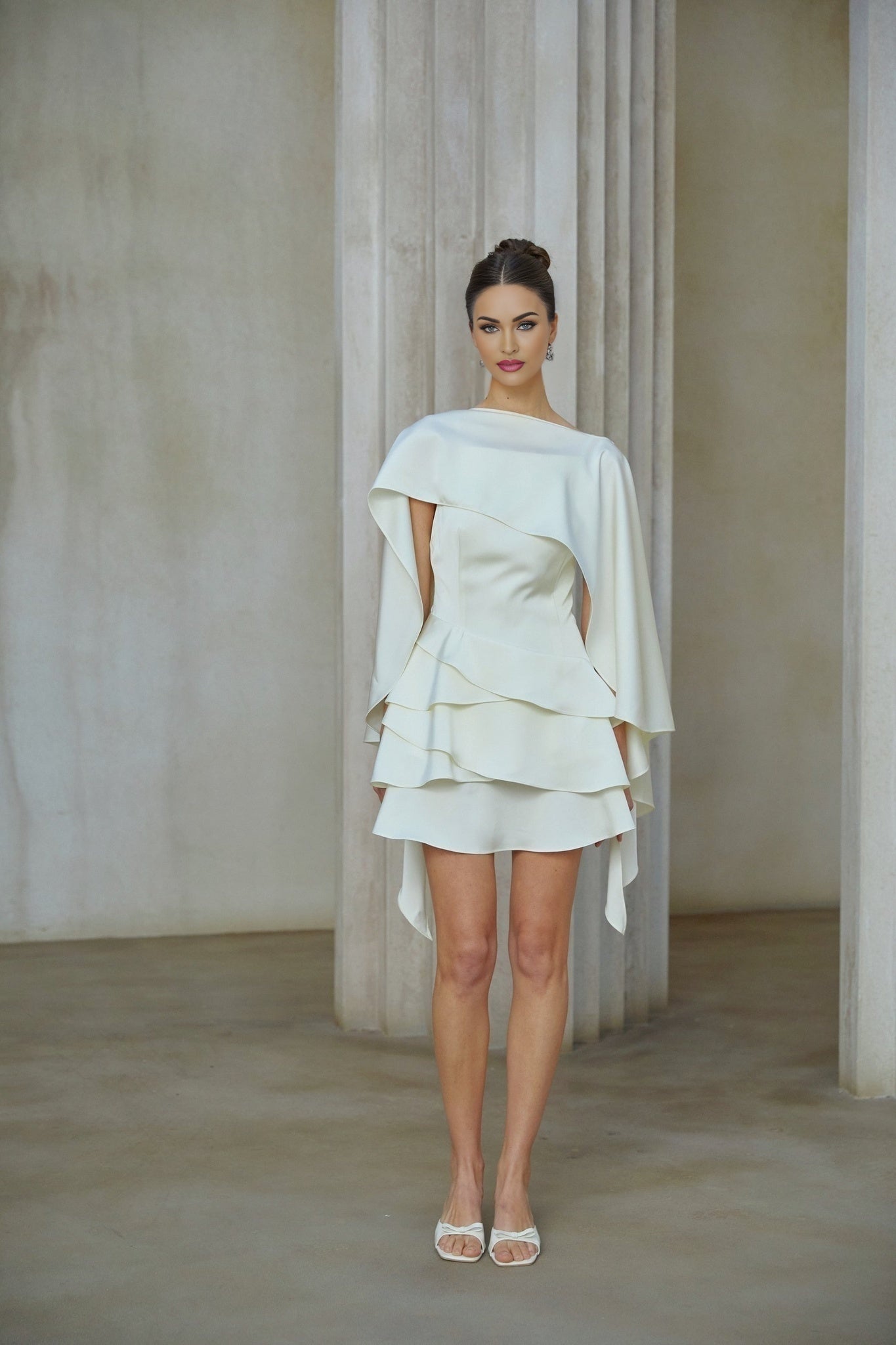 A white satin mini dress with ruffled details and a shawl panel feature on a model against a plain background