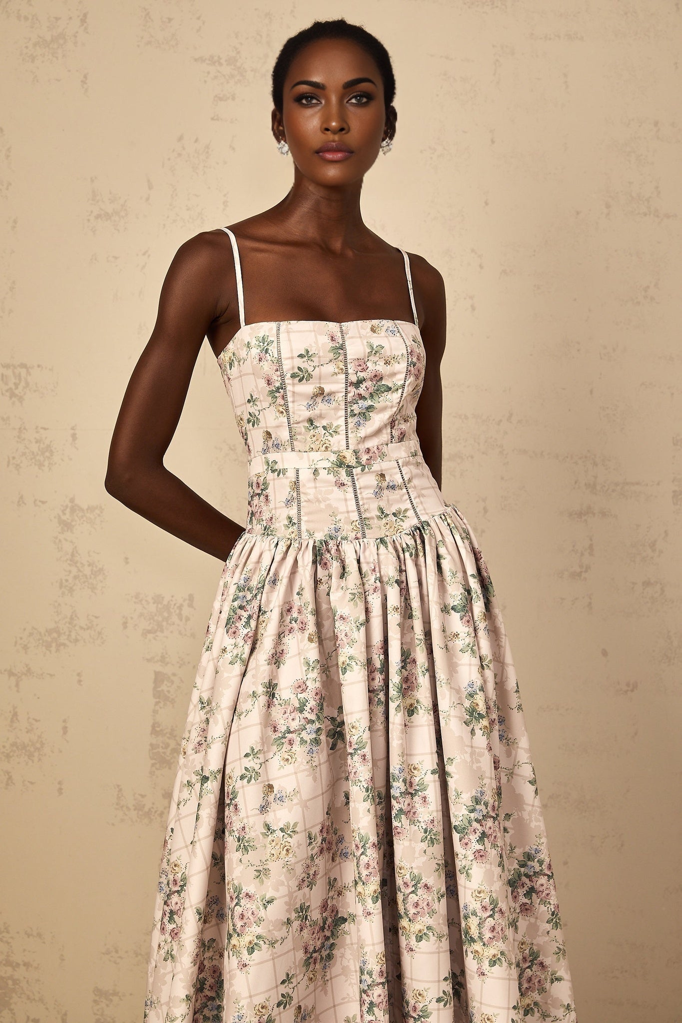 A woman wearing a beige midi dress with a delicate floral print pattern standing in a relaxed pose against a plain background