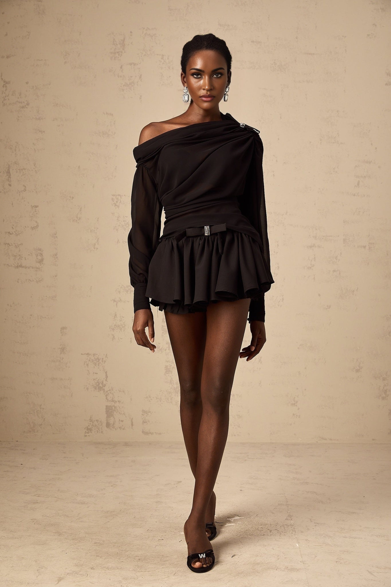 A woman wearing a black asymmetric tulle blouse and matching skirt set The top features a one shoulder design with ruffled details The skirt is flowy and made of tulle fabric The outfit is elegant and suitable for formal occasions