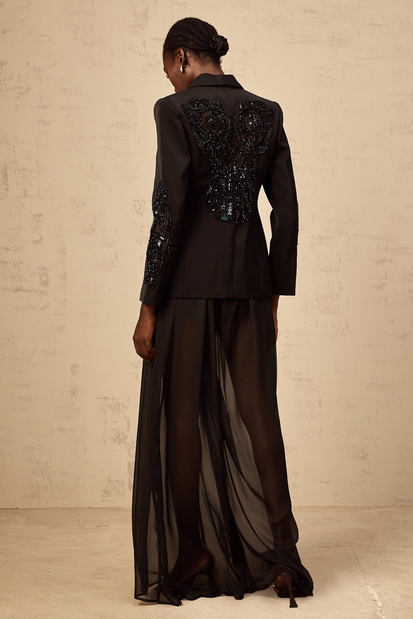 A woman wearing a black blazer and trousers set with sheer tulle panels and embellishments standing in a neutral pose