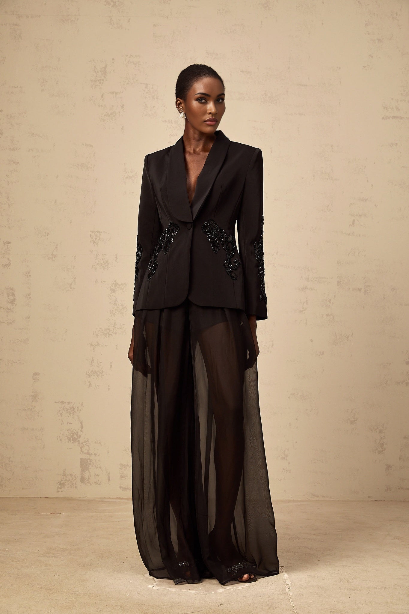 A woman wearing a black blazer and trousers set with sheer tulle panels and embellishments standing in a neutral pose