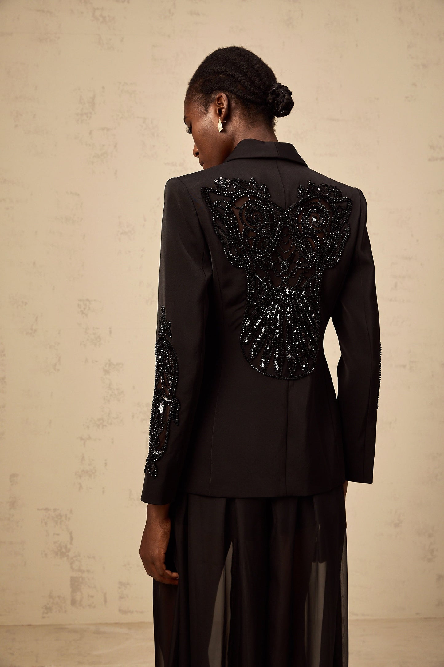 A woman wearing a black blazer and trousers set with sheer tulle panels and embellishments standing in a neutral pose