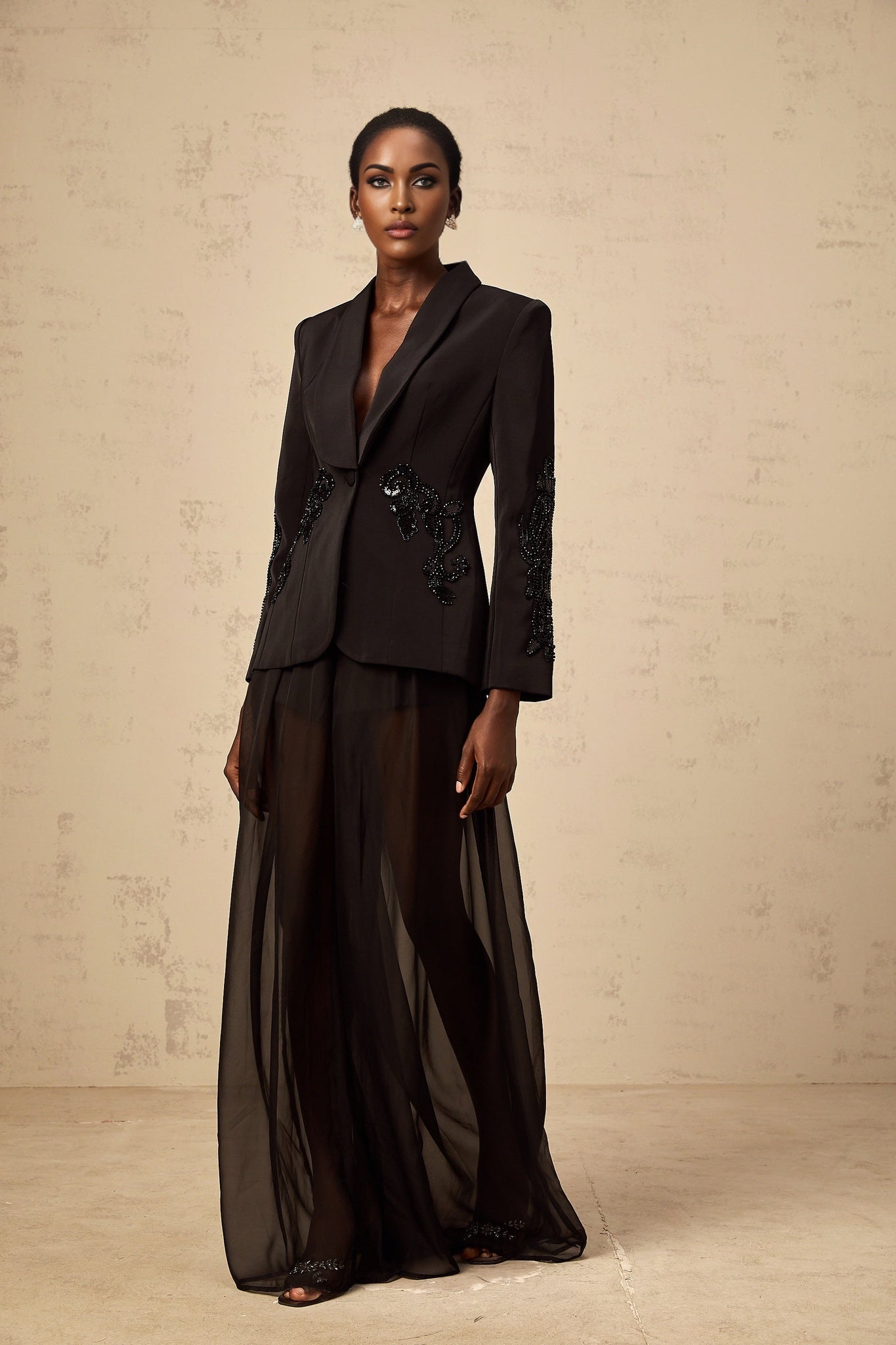 A woman wearing a black blazer and trousers set with sheer tulle panels and embellishments standing in a neutral pose