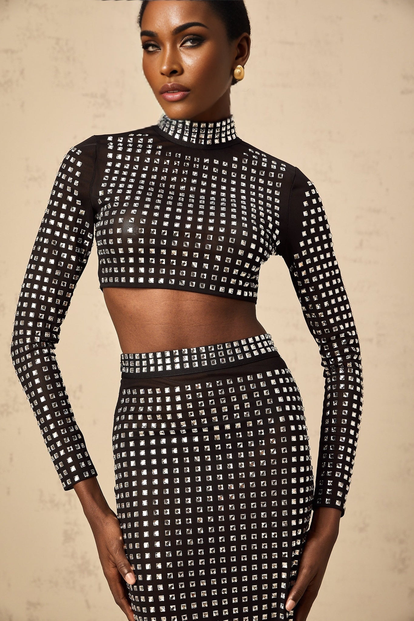 A woman wearing a black matching set consisting of a sleeveless mesh top and skirt adorned with sparkling rhinestones The top features a high neck while the skirt has a figure hugging silhouette Both pieces are made of sheer mesh fabric embellished with a pattern of glittering black rhinestones