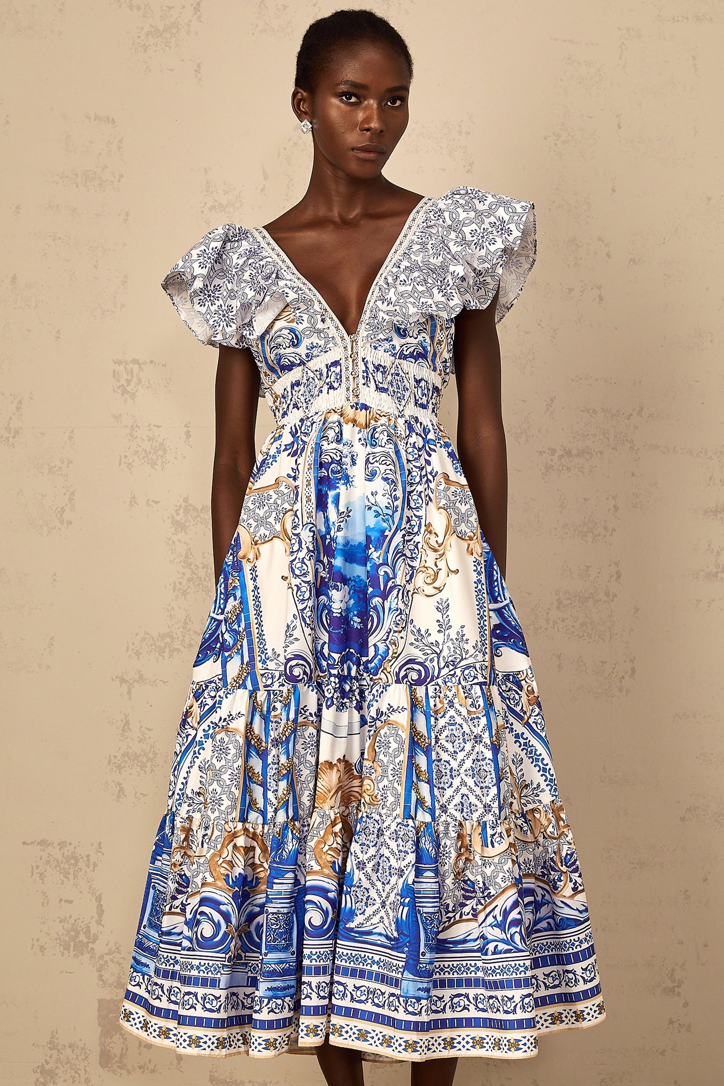 A woman wearing a blue midi dress with a baroque print pattern and tiered design standing in a neutral background