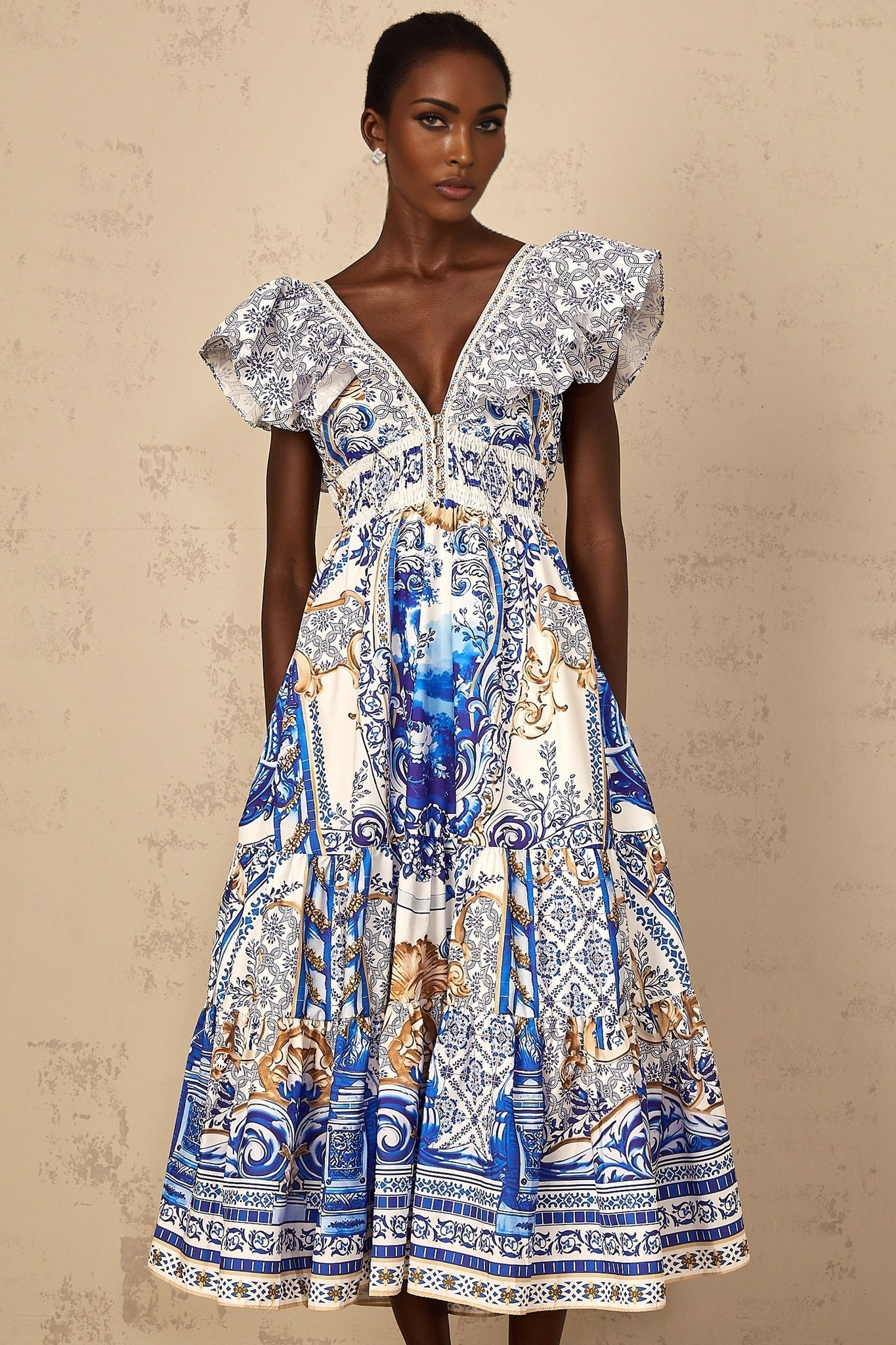A woman wearing a blue midi dress with a baroque print pattern and tiered design standing in a neutral background