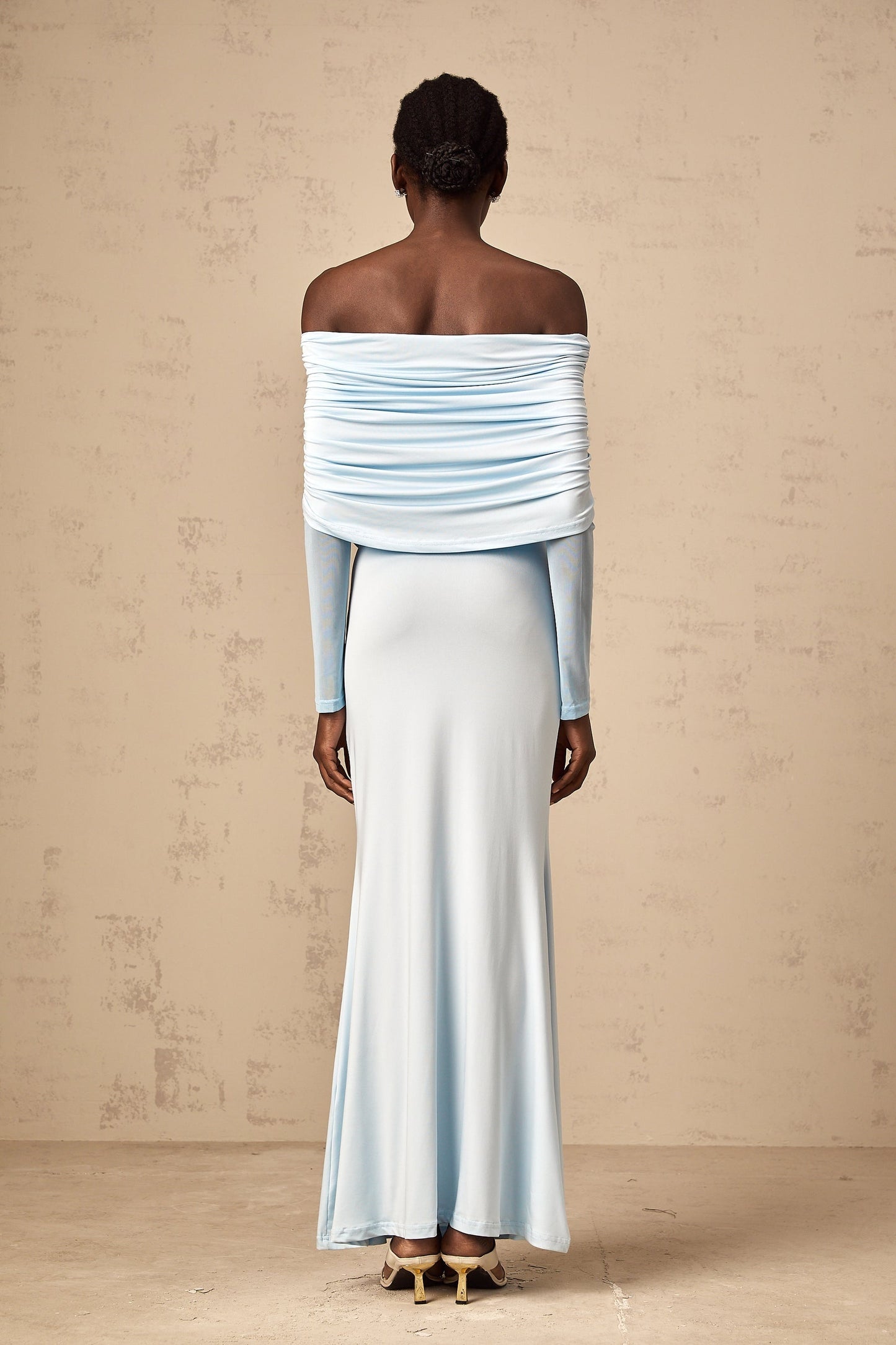A woman wearing a blue offshoulder maxi dress with ruched detailing standing against a plain background