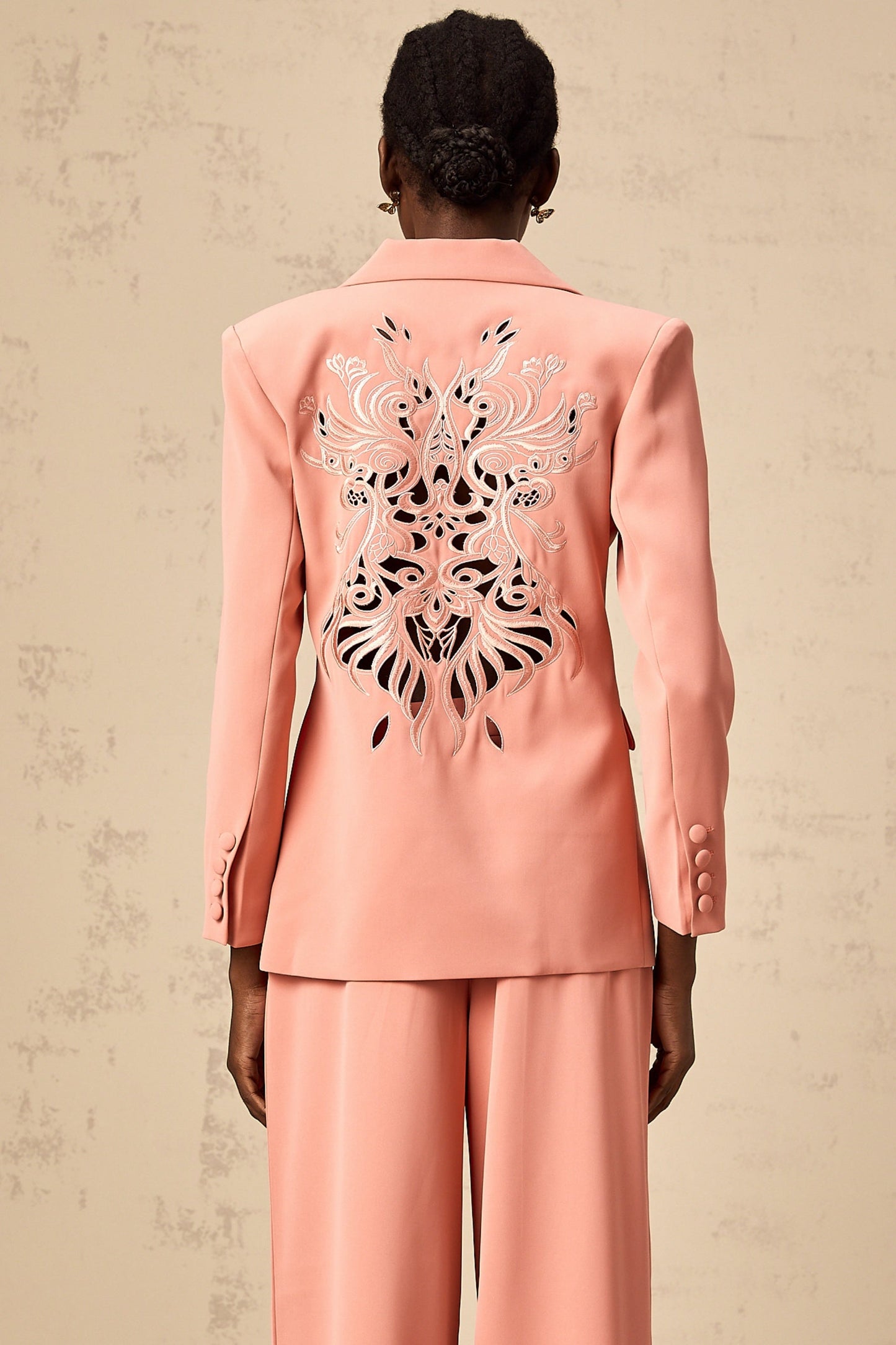 A woman wearing a bright pink matching set consisting of a blazer with cut out details and embroidery paired with tailored trousers