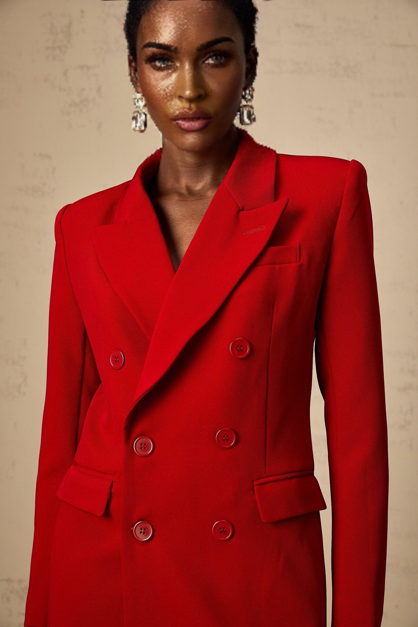 A woman wearing a bright red doublebreasted blazer with peaked lapels and long sleeves standing against a plain background