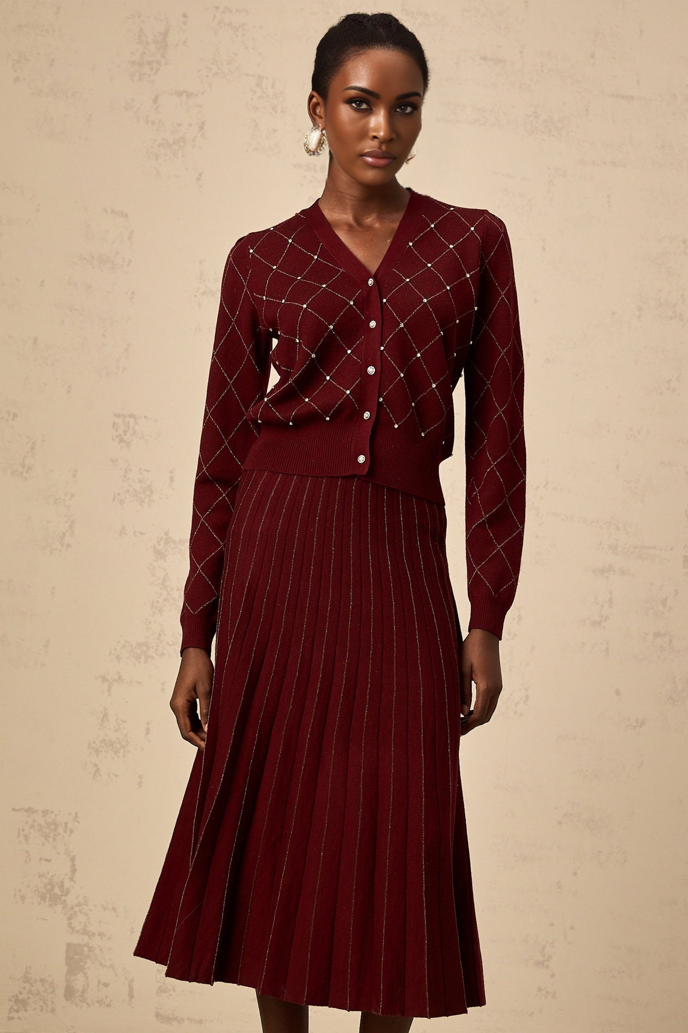 A woman wearing a bright red matching set consisting of a cardigan and skirt The cardigan features an argyle pattern with embellishments and the skirt is knitted in a coordinating style Both pieces have intarsia knit detailing The outfit is styled as a fashionable two piece ensemble