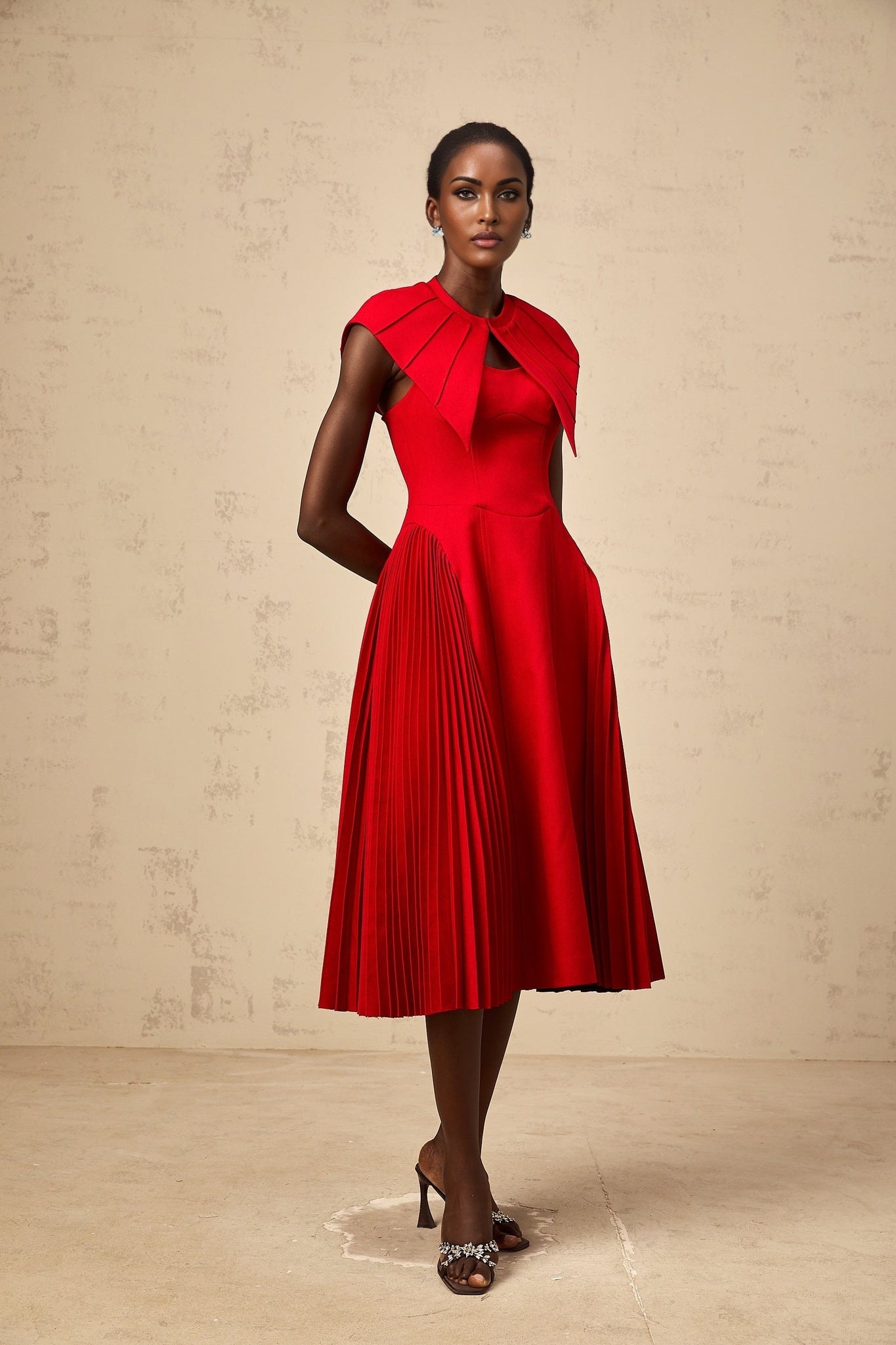 A woman wearing a bright red midi dress with a capelike design and pleated skirt standing against a plain background