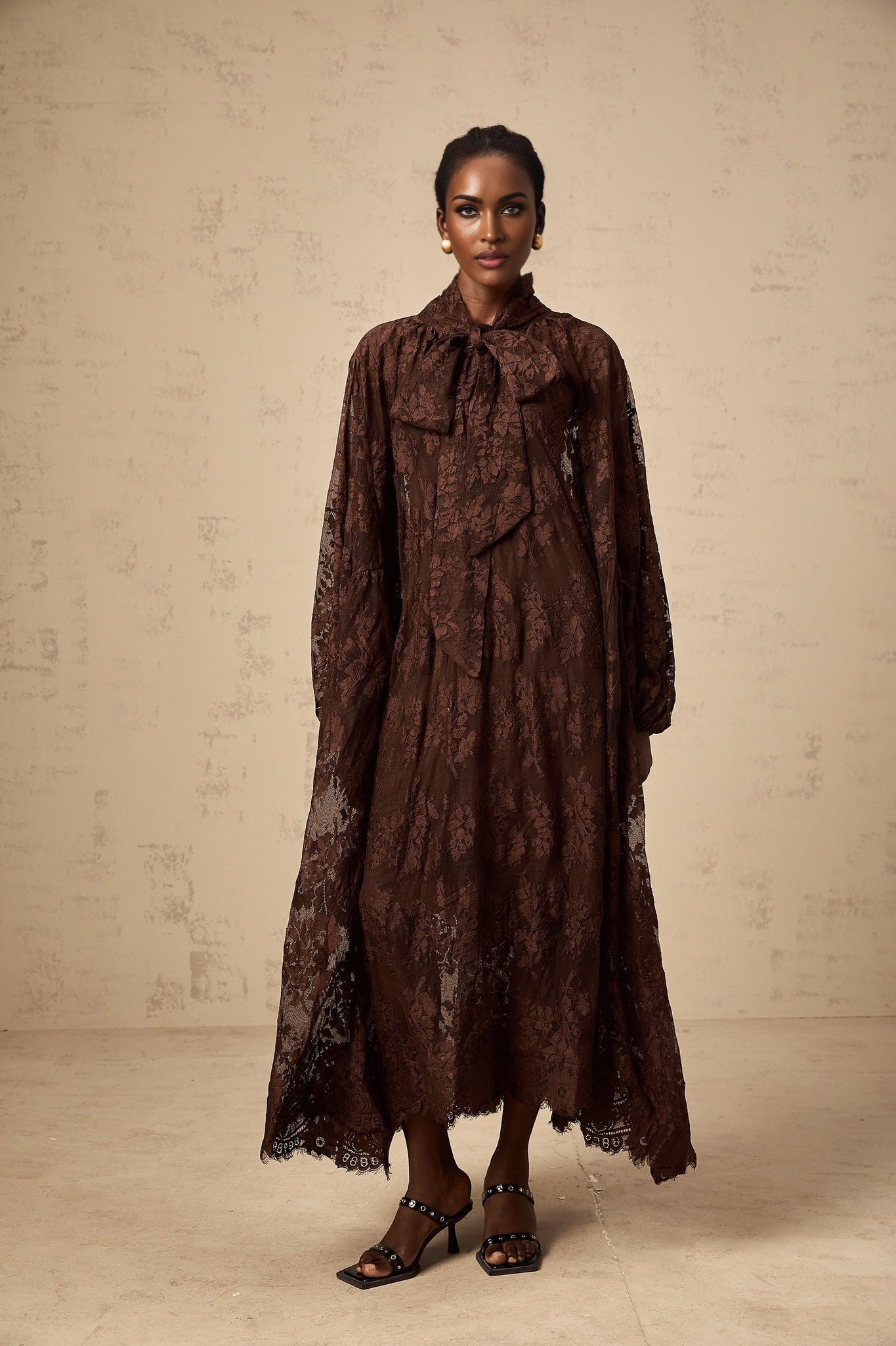 A woman wearing a brown flared midi dress made of chantilly lace named Sylvie standing in a stylish pose