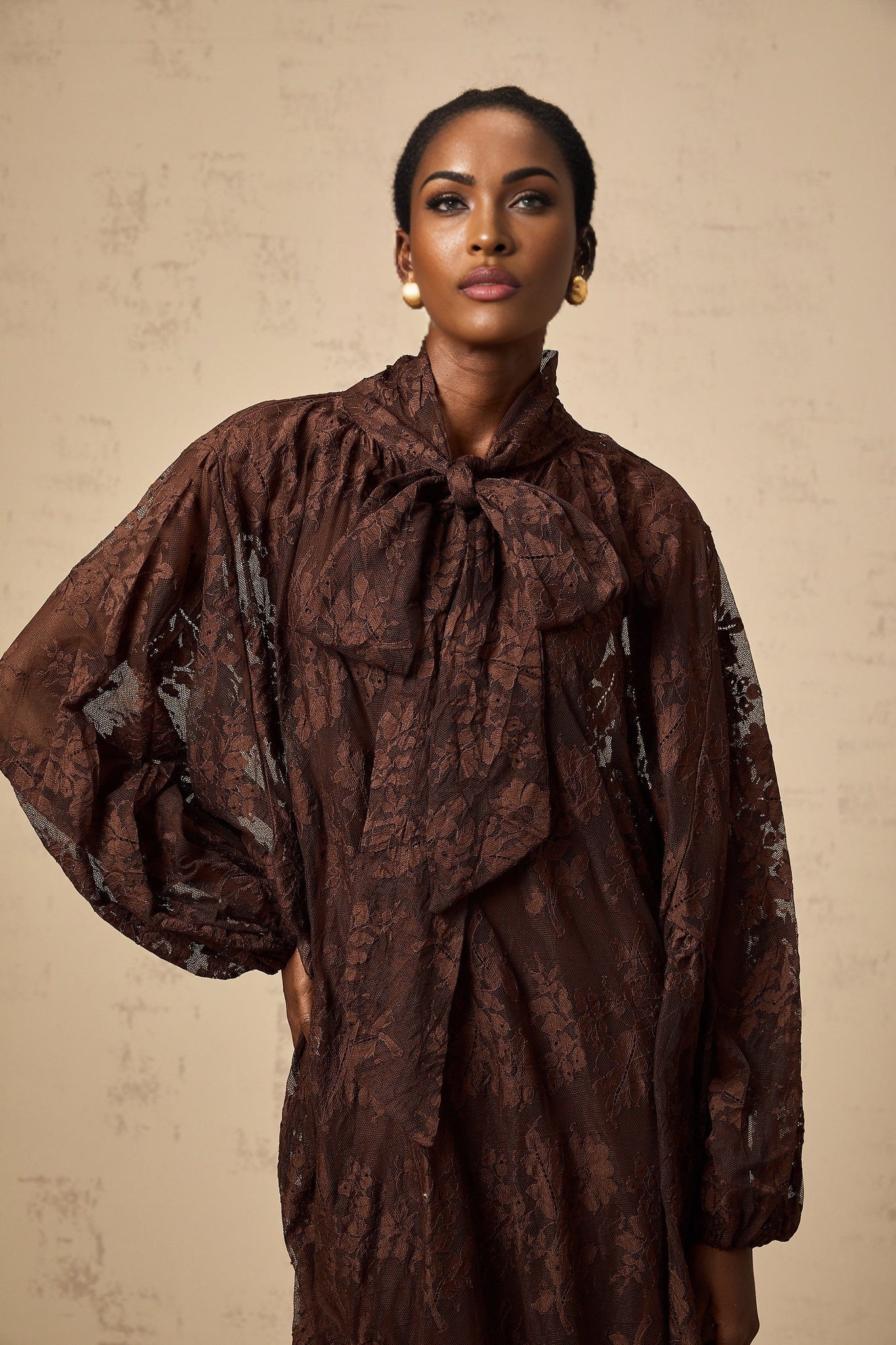 A woman wearing a brown flared midi dress made of chantilly lace named Sylvie The dress features a fitted bodice and a flowy skirt that falls just below the knee