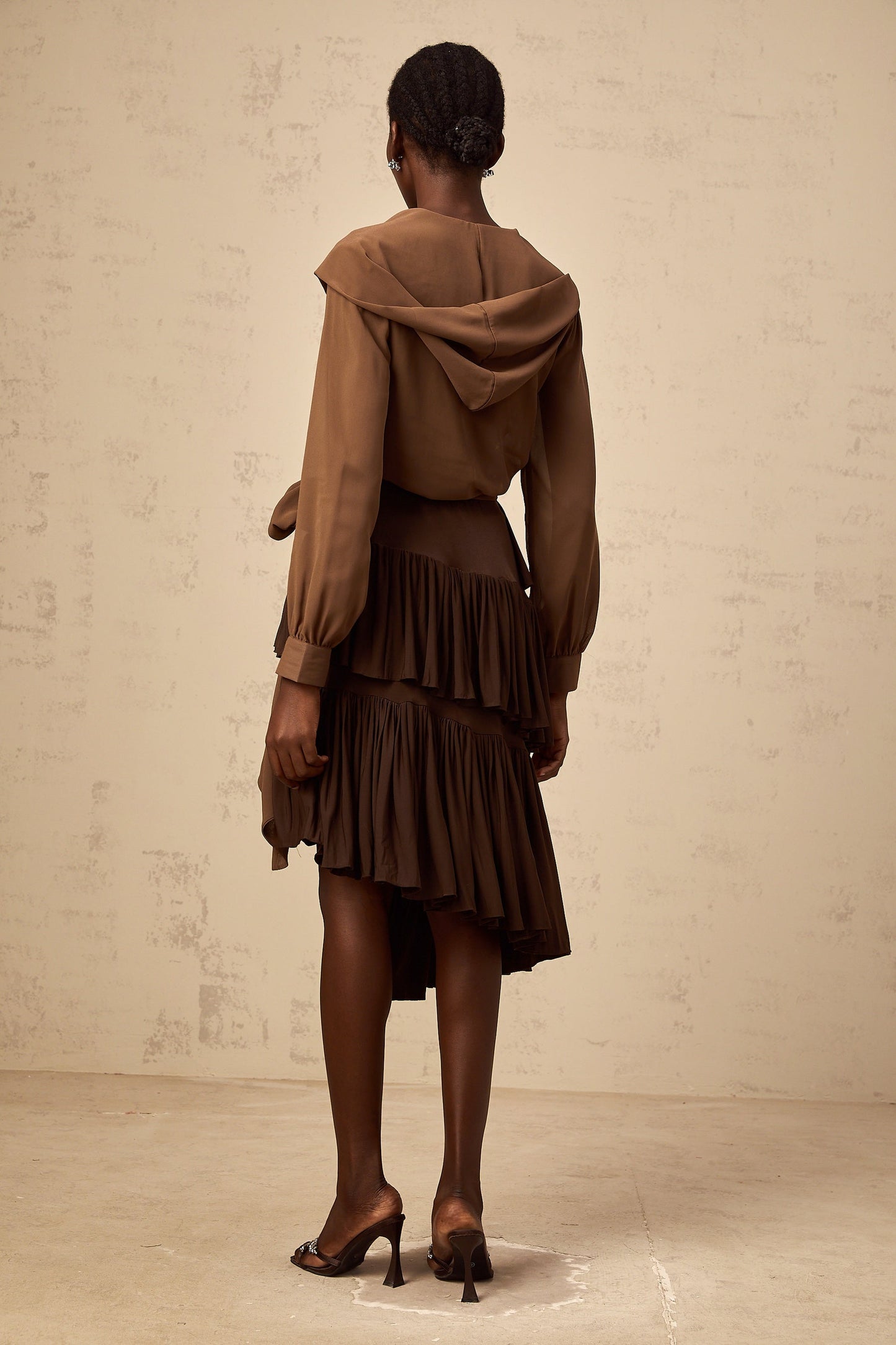 A woman wearing a brown hooded wrap top with semi sheer fabric The top has long sleeves and drapes loosely around the body
