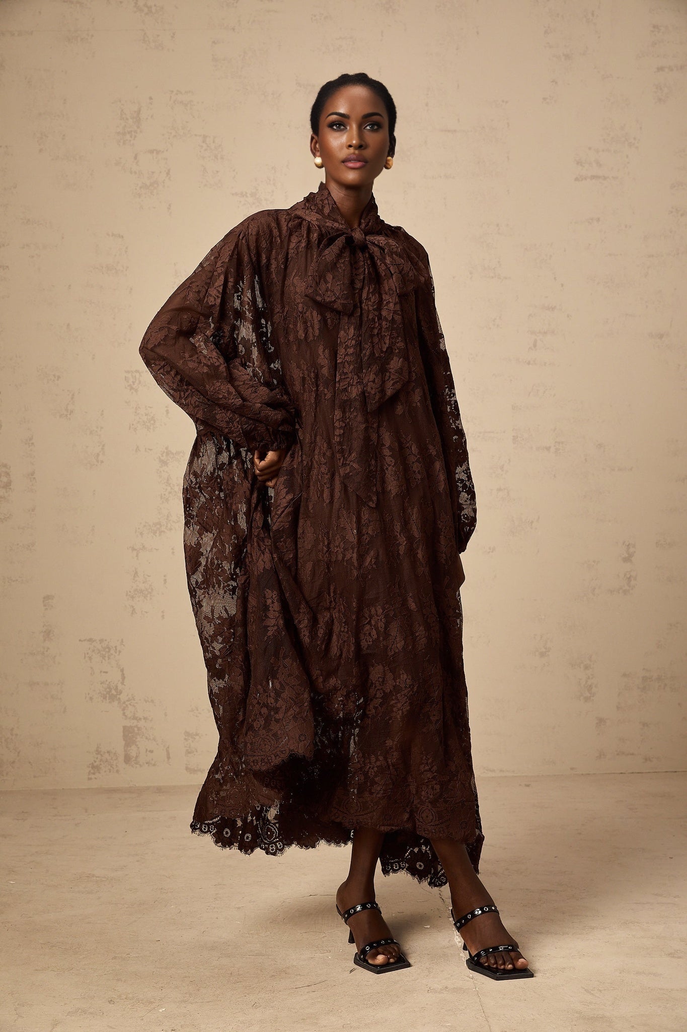 A woman wearing a brown lace midi dress with a flared skirt and delicate chantilly lace pattern The dress has a fitted bodice and falls to mid calf length
