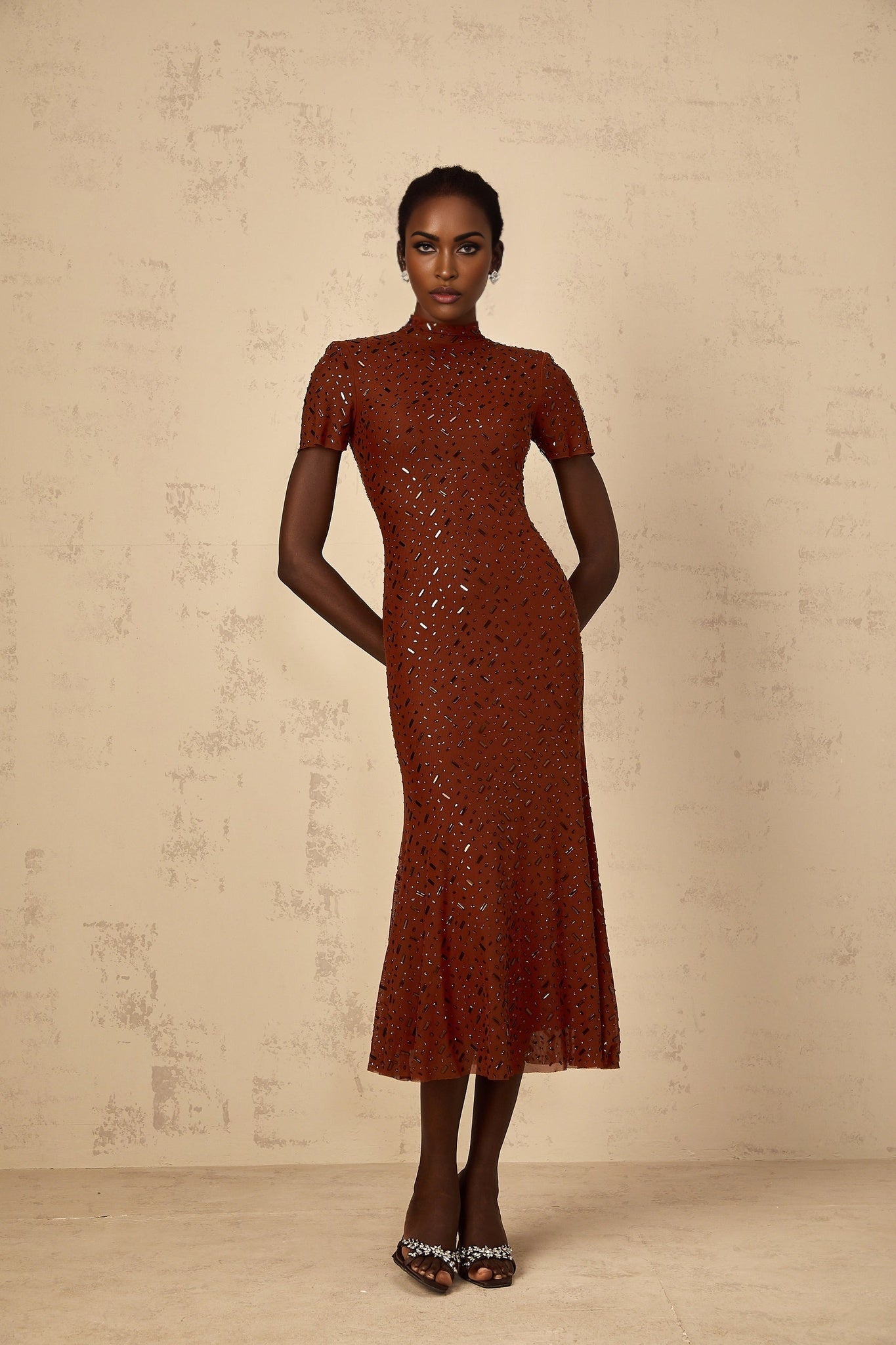 A woman wearing a brown midi dress with a flared tulle skirt and embellished details standing against a plain background