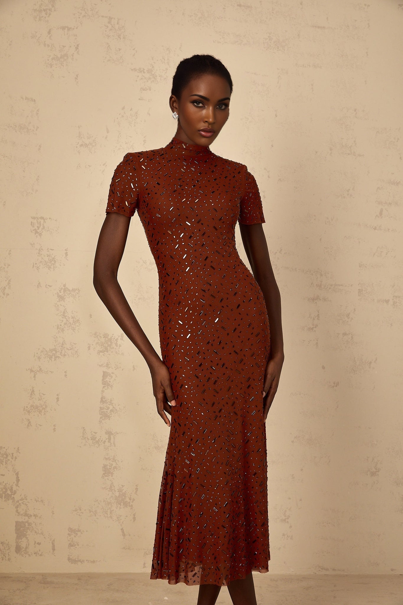 A woman wearing a brown midi dress with a flared tulle skirt and embellished details standing in a neutral background