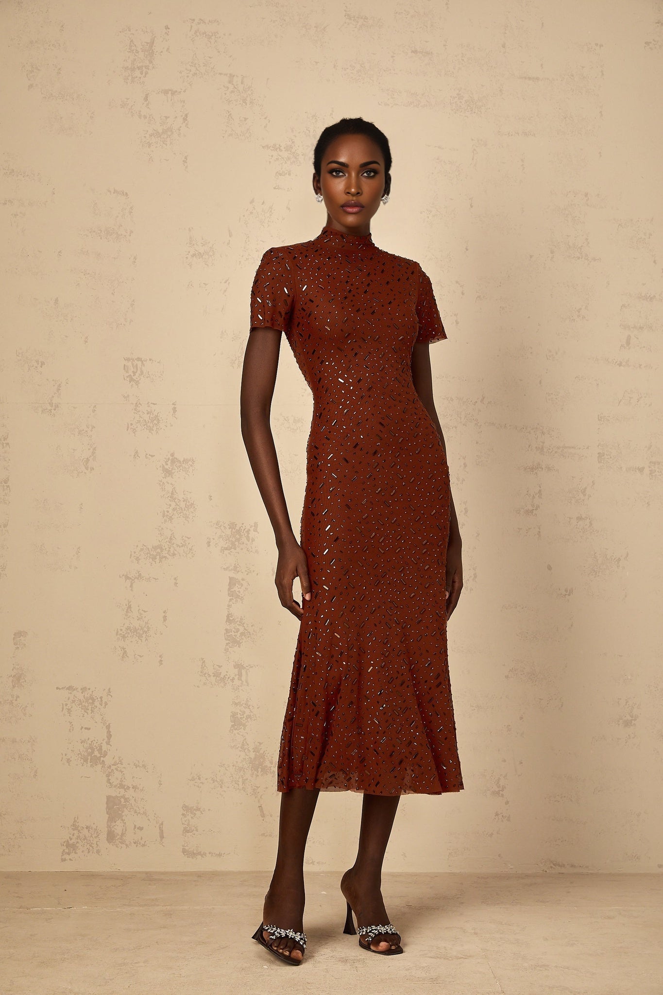 A woman wearing a brown midi dress with a flared tulle skirt and embellished details standing in a neutral pose