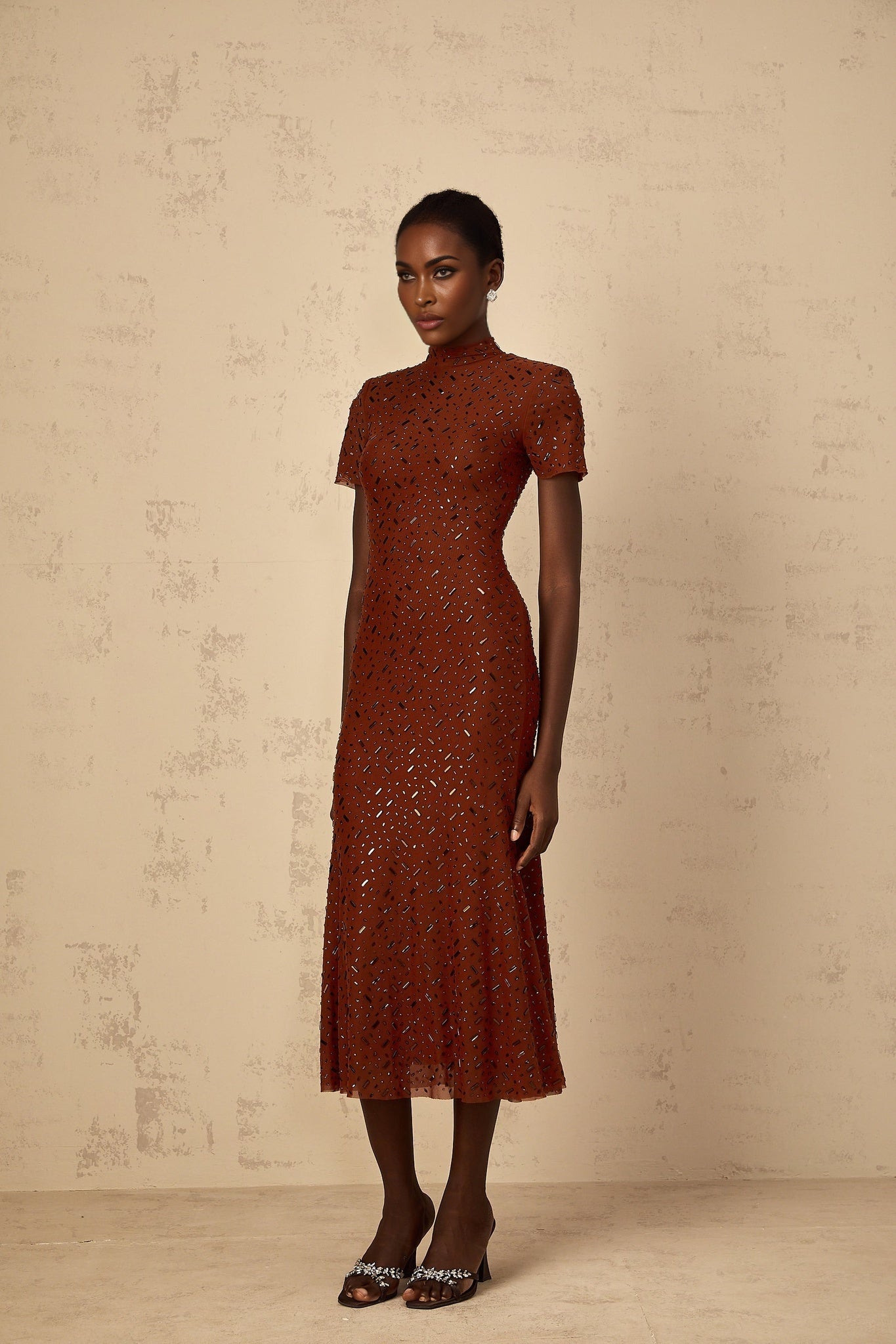 A woman wearing a brown midi dress with a flared tulle skirt and embellished details standing in a neutral pose