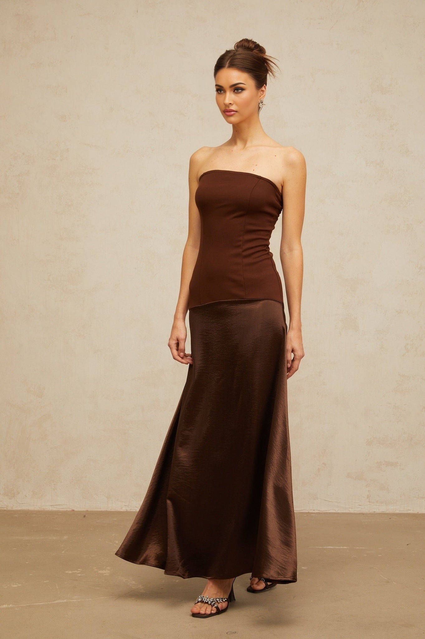 A woman wearing a brown offshoulder maxi dress with satin panels standing against a plain background