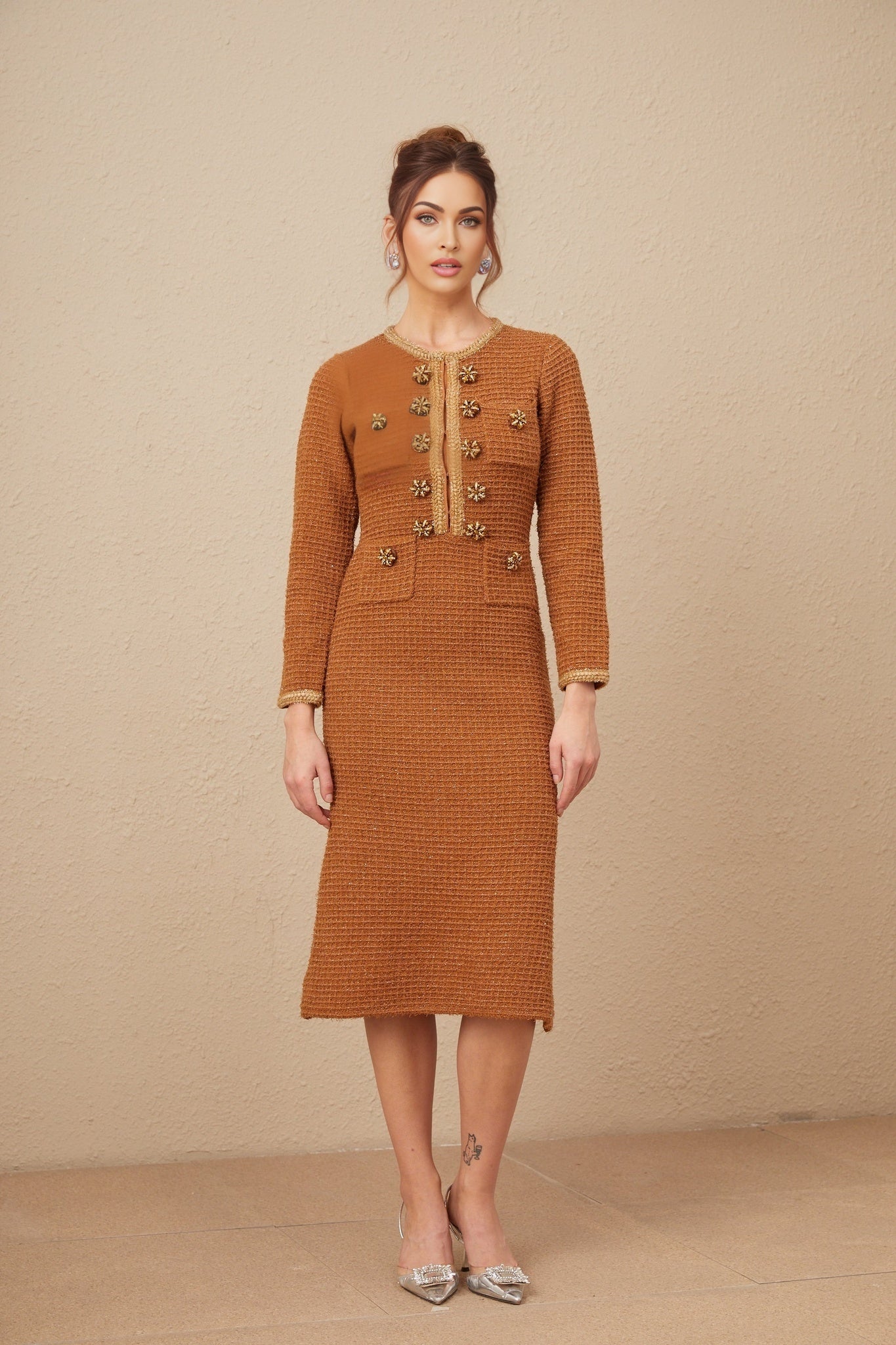 A woman wearing a brown tweed dress with braided edges and a fitted silhouette standing against a plain background