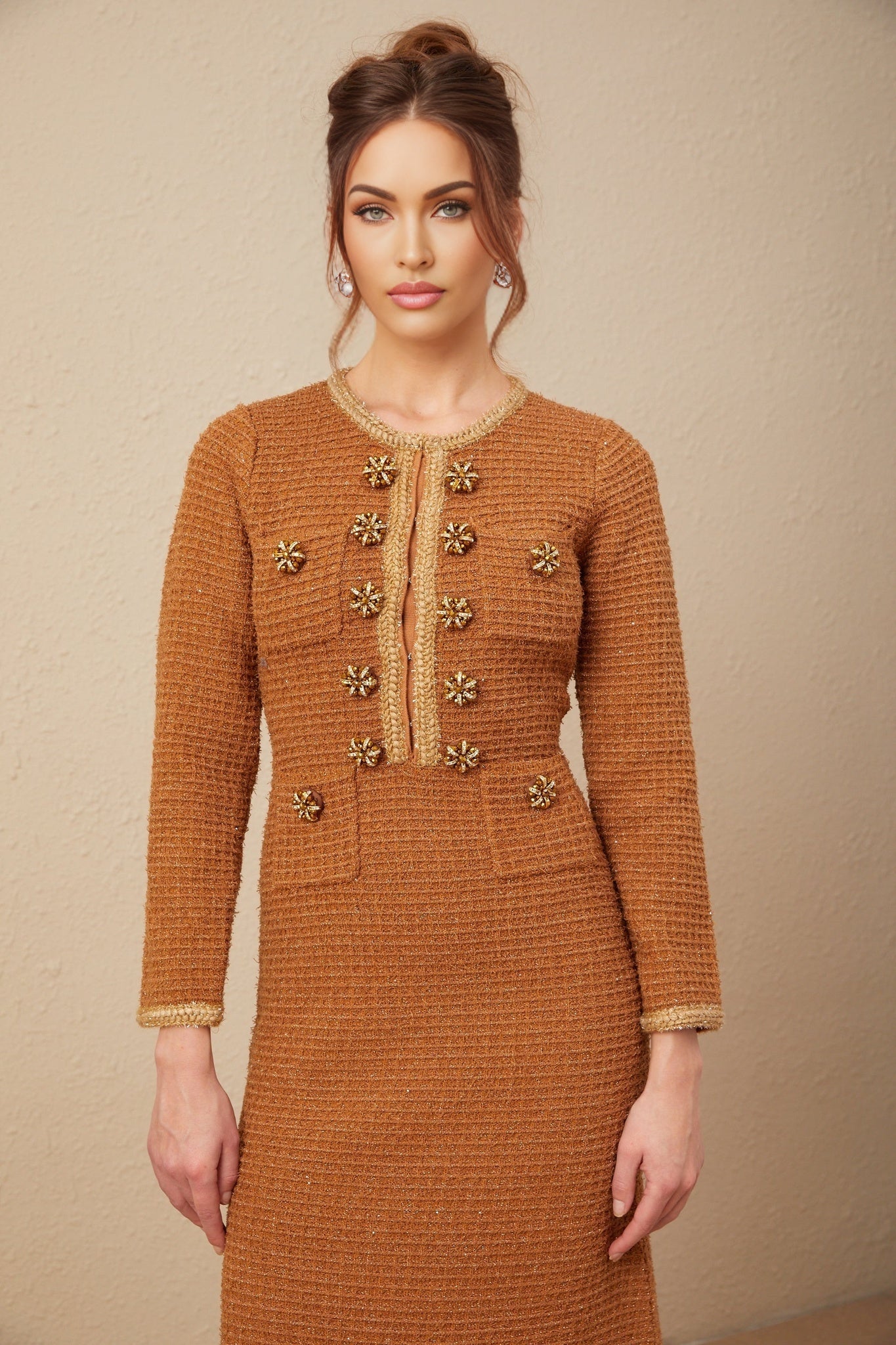 A woman wearing a brown tweed dress with braided edges standing against a plain background
