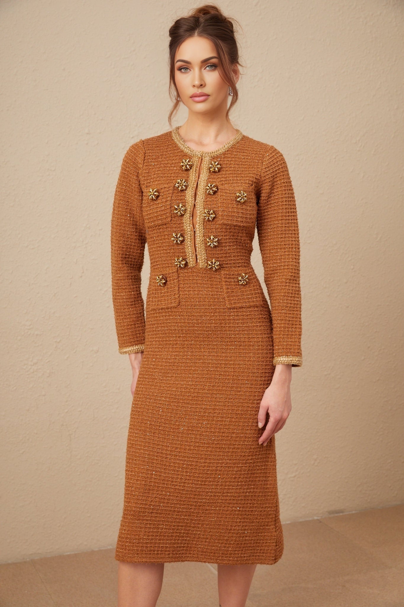 A woman wearing a brown tweed dress with braided edges standing in a neutral background