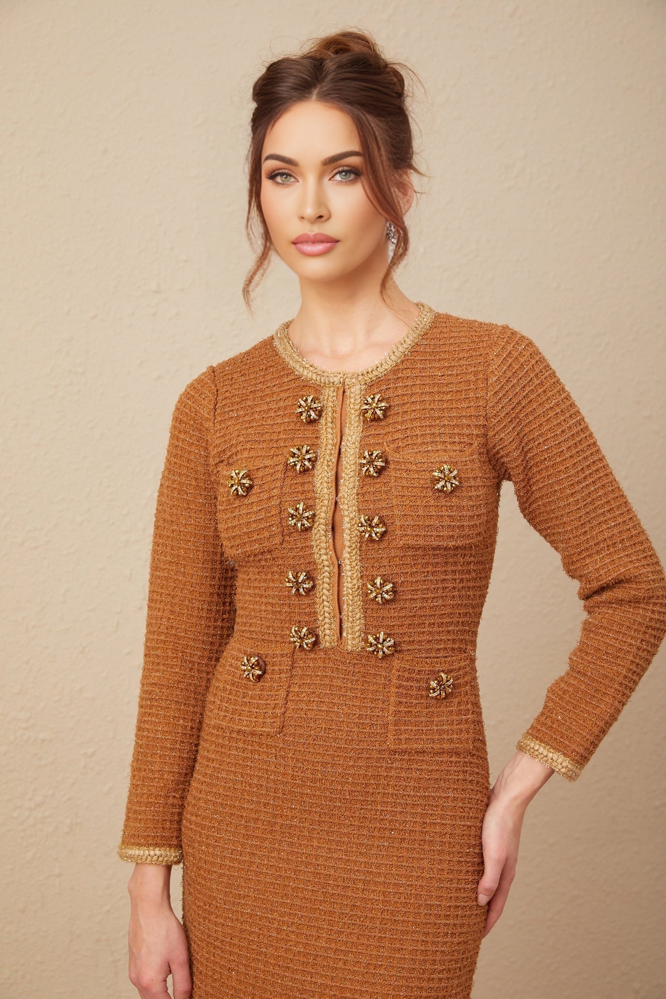 A woman wearing a brown tweed dress with braided edges standing in a neutral pose against a plain background