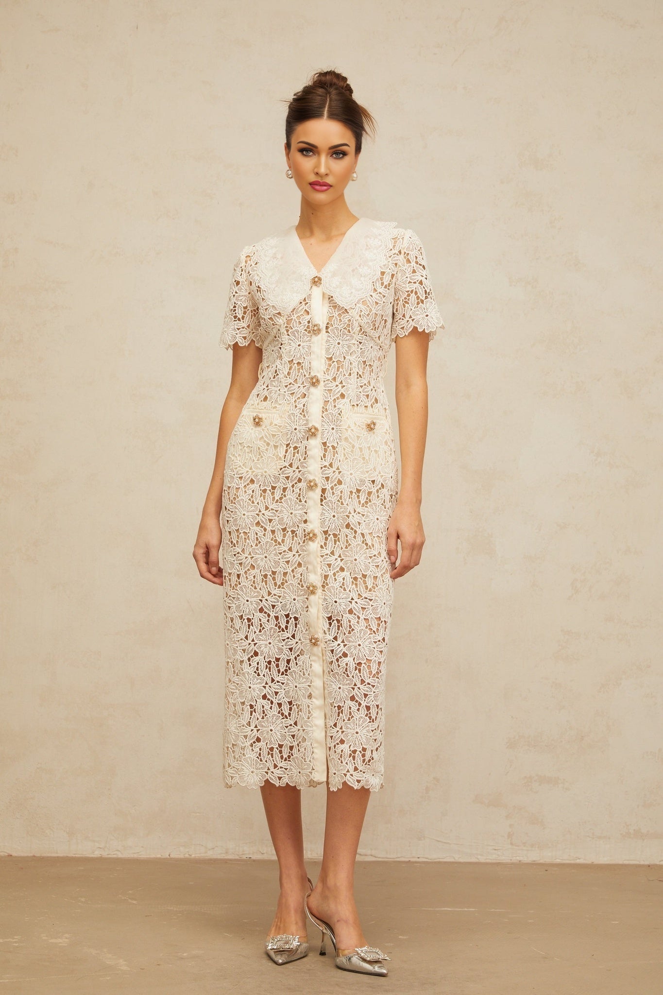 A woman wearing a cream colored lace midi dress with off the shoulder sleeves standing in a neutral background
