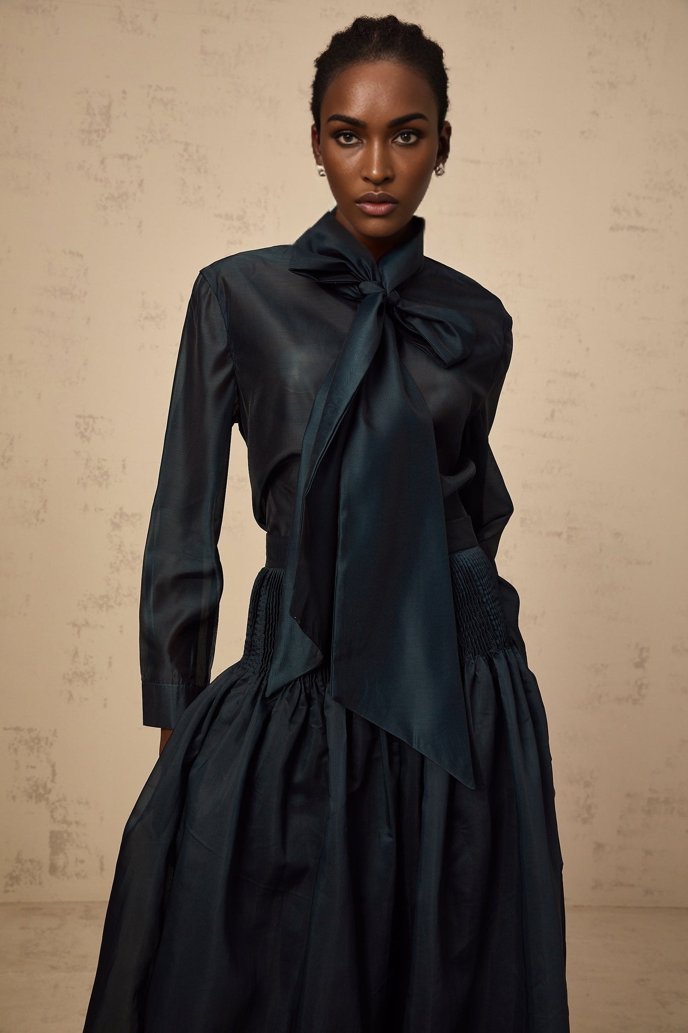 A woman wearing a dark green matching set consisting of a long sleeve shirt with a ribbon tie neck and a coordinating skirt