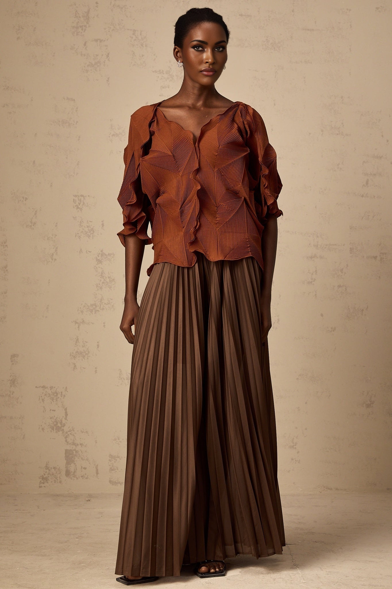 A woman wearing a demi brown matching set consisting of a creased top and skirt standing against a plain background