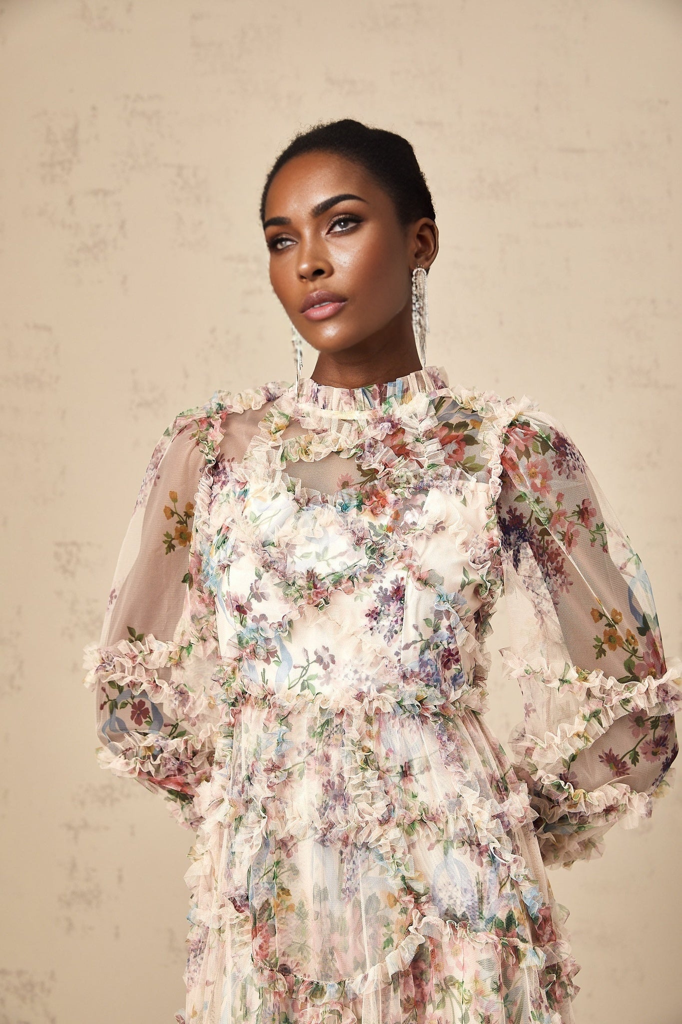 A woman wearing a floral patterned midi dress with ruffled details and a tulle overlay standing against a plain background