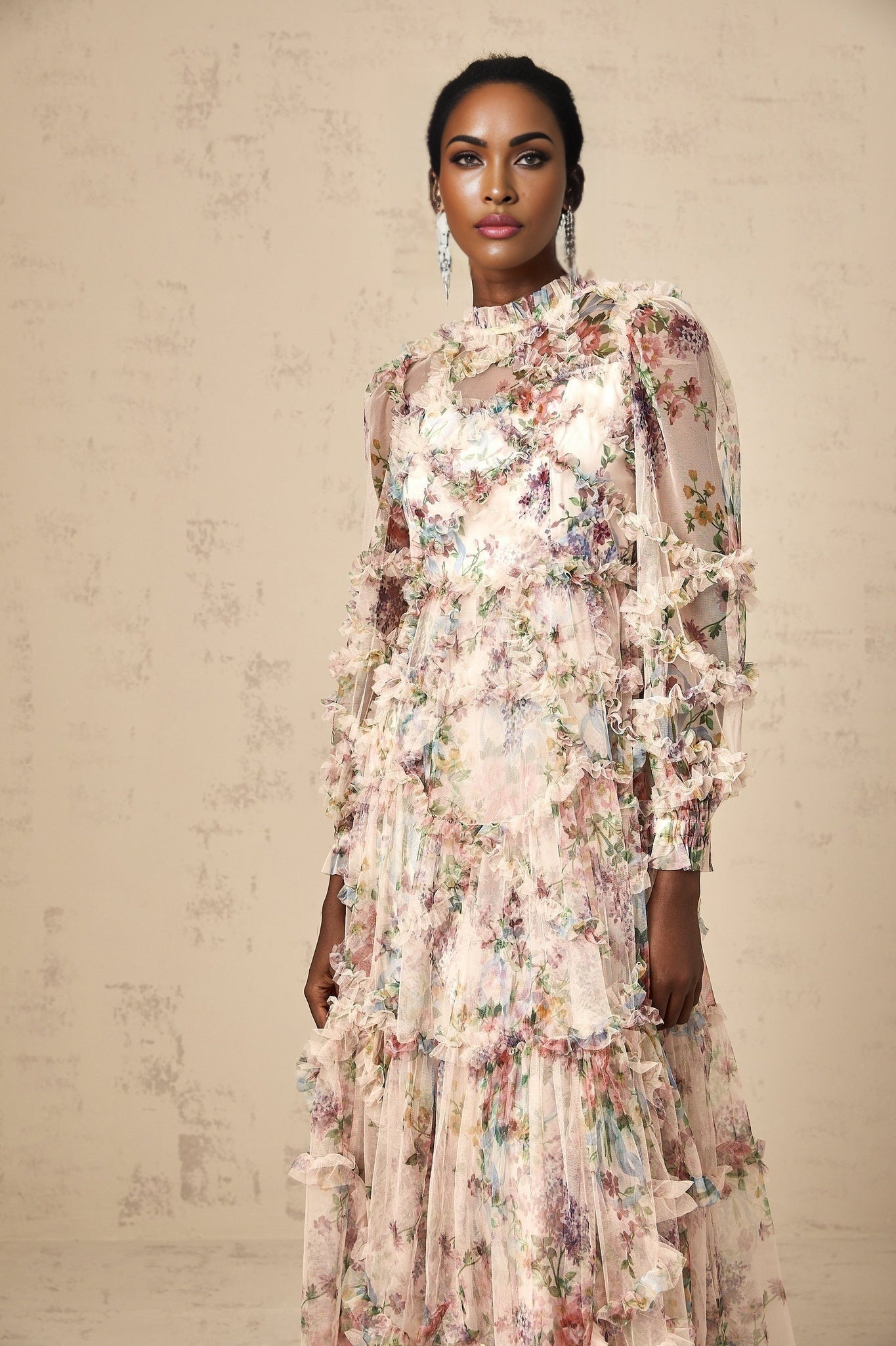 A woman wearing a floral patterned midi dress with ruffled details and a tulle overlay standing against a plain background