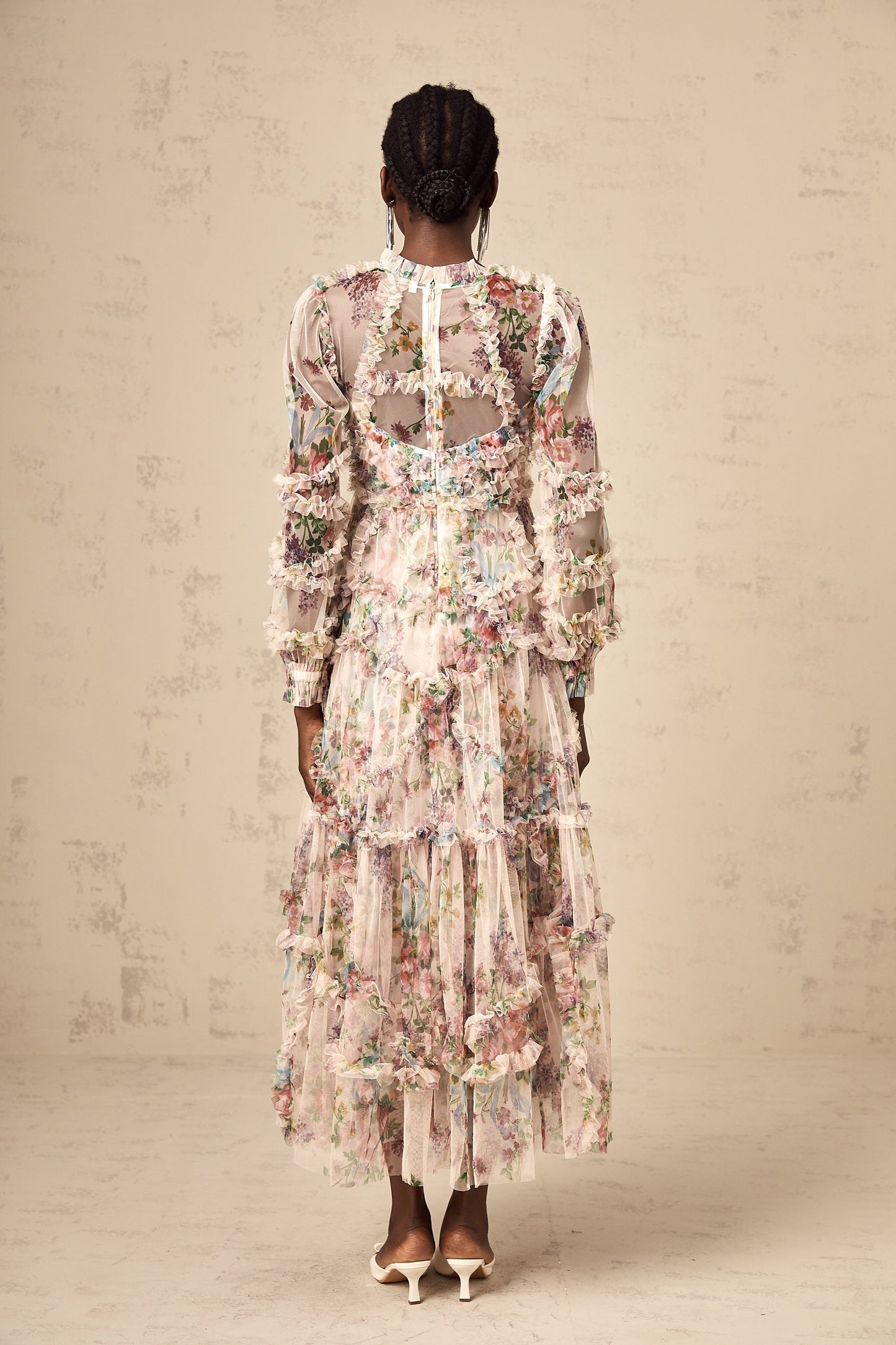 A woman wearing a floral patterned midi dress with ruffled details and a tulle overlay standing against a plain background