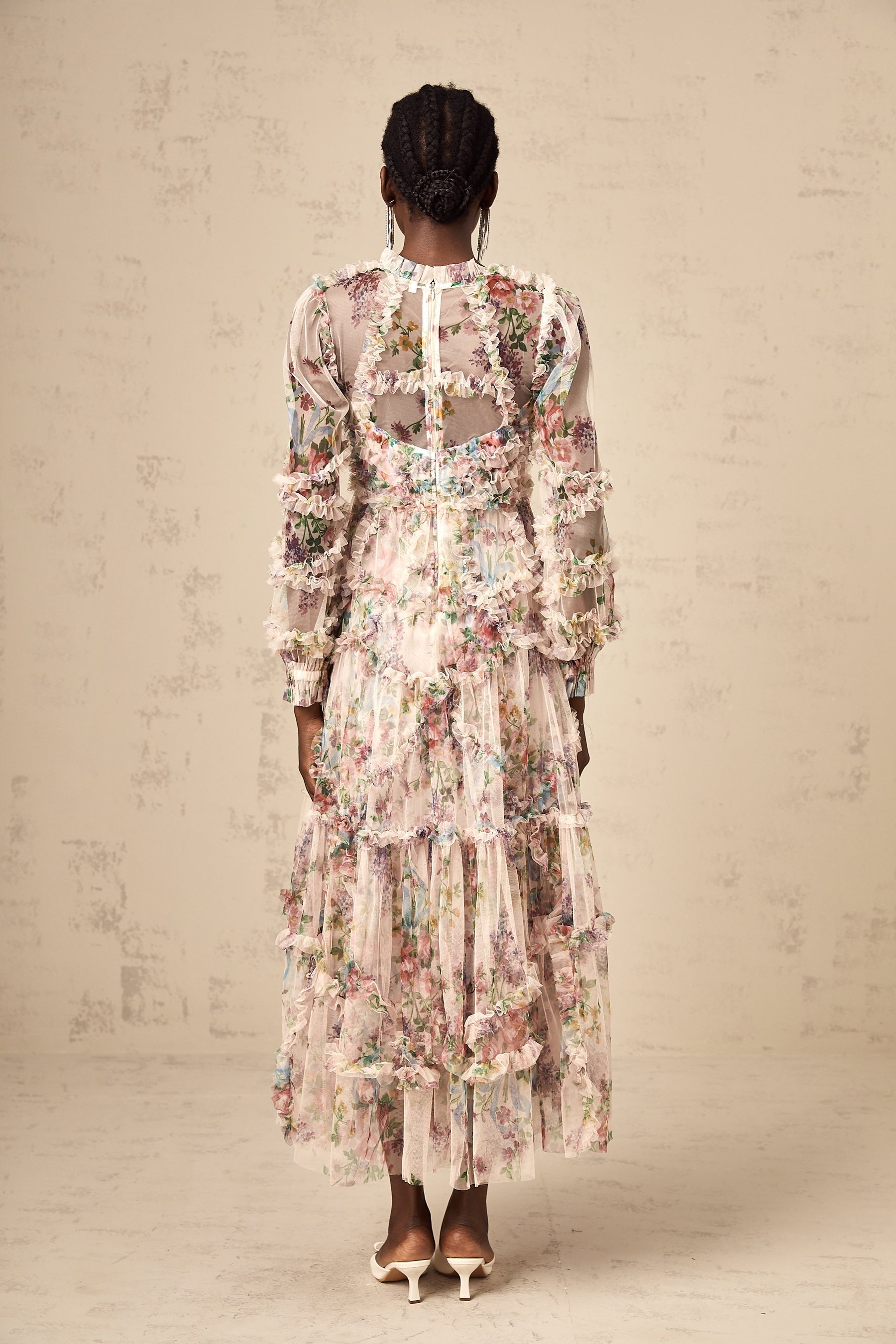 A woman wearing a floral patterned midi dress with ruffled details and a tulle overlay standing against a plain background