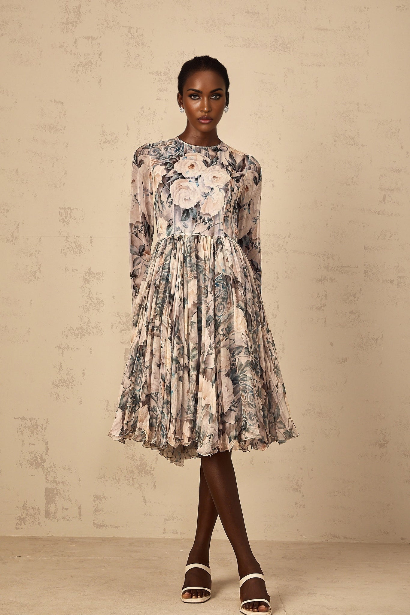 A woman wearing a floral print chiffon midi dress with pleated details standing in a relaxed pose against a plain background