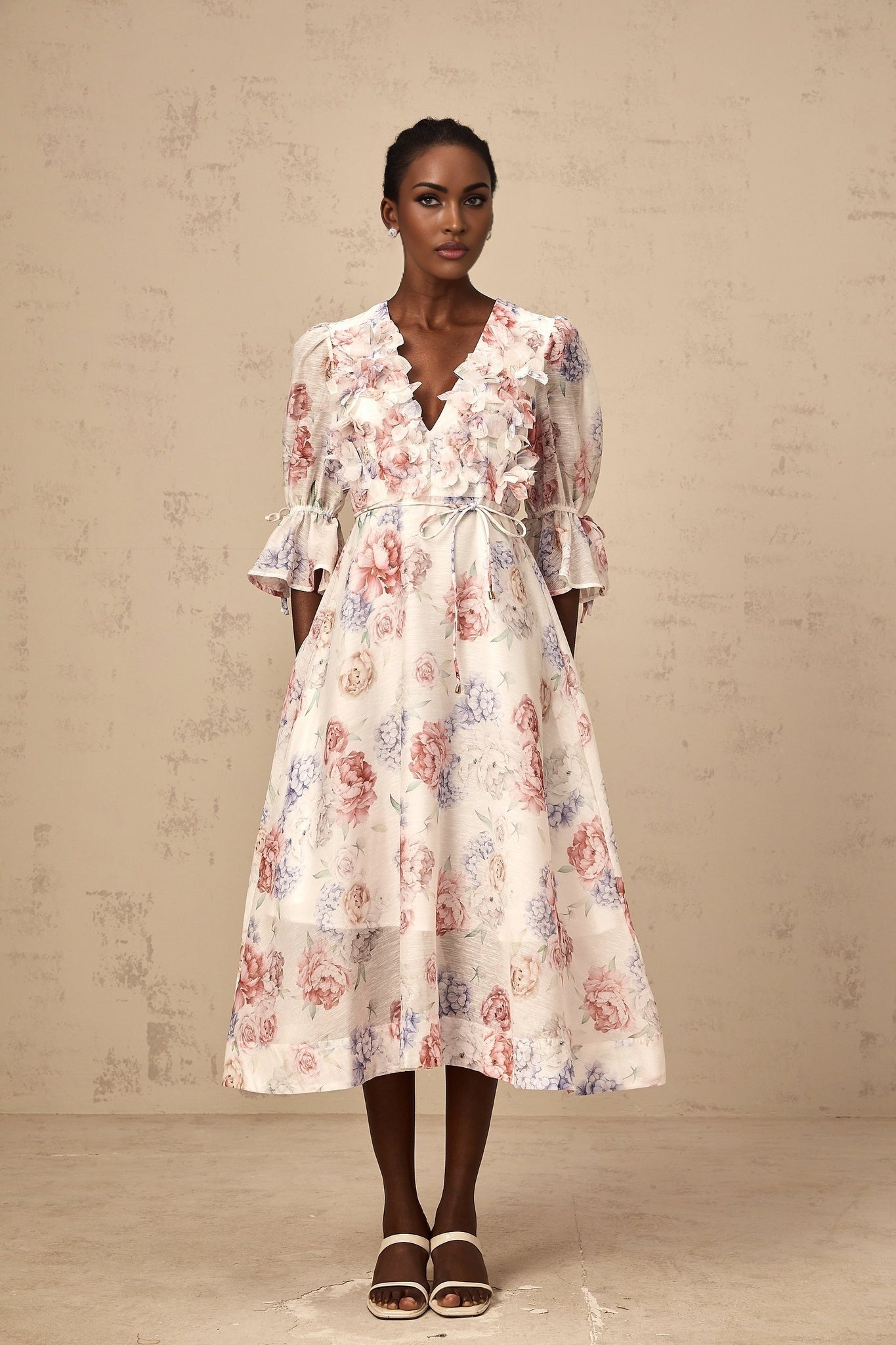 A woman wearing a floral print midi dress with three dimensional petal details on a light colored background
