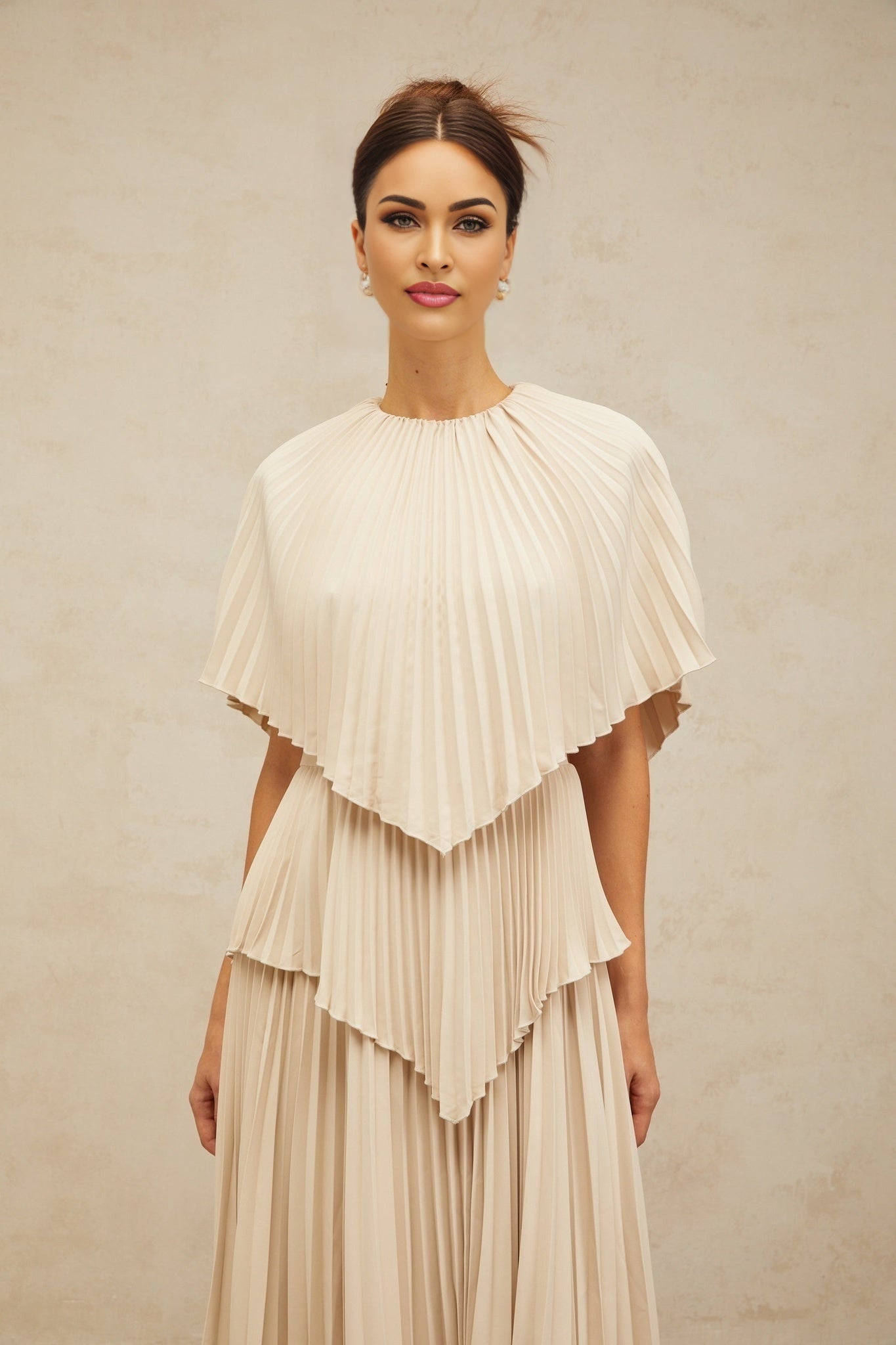 A woman wearing a flowing neutralcolored midi dress with cape detailing and pleated skirt standing in a relaxed pose