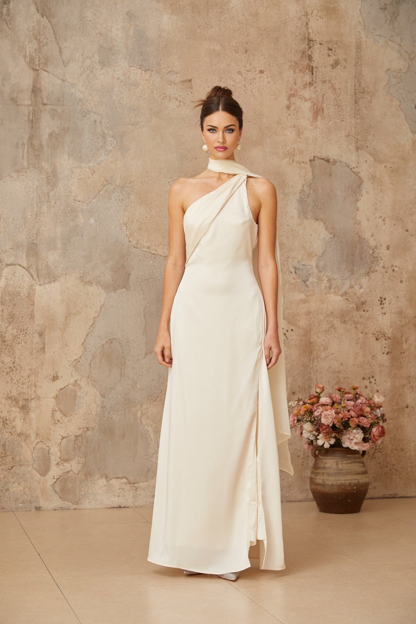 A woman wearing a flowing white satin maxi dress with a scarflike design detail at the neckline standing against a plain background
