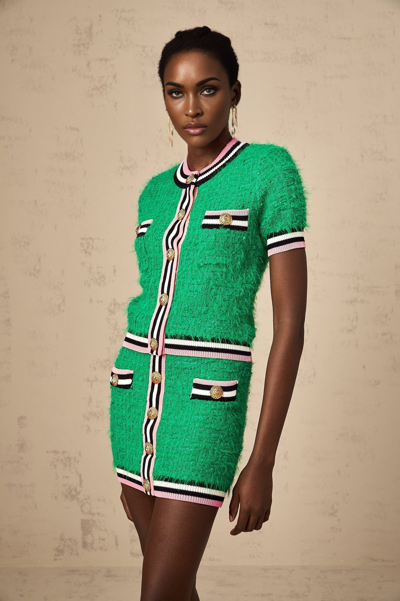 A woman wearing a green knitted two piece outfit consisting of a top and skirt set The top has long sleeves and the skirt is knee length The model is standing against a plain background