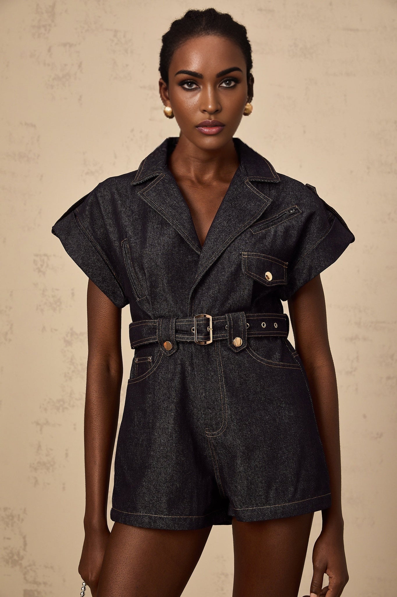 A woman wearing a light blue denim playsuit with a belted waist standing against a plain background