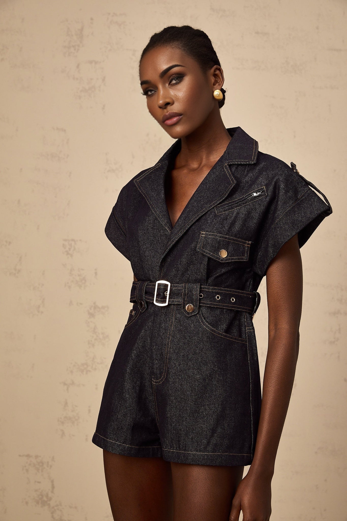 A woman wearing a light blue denim playsuit with short sleeves and a belted waist standing in a relaxed pose