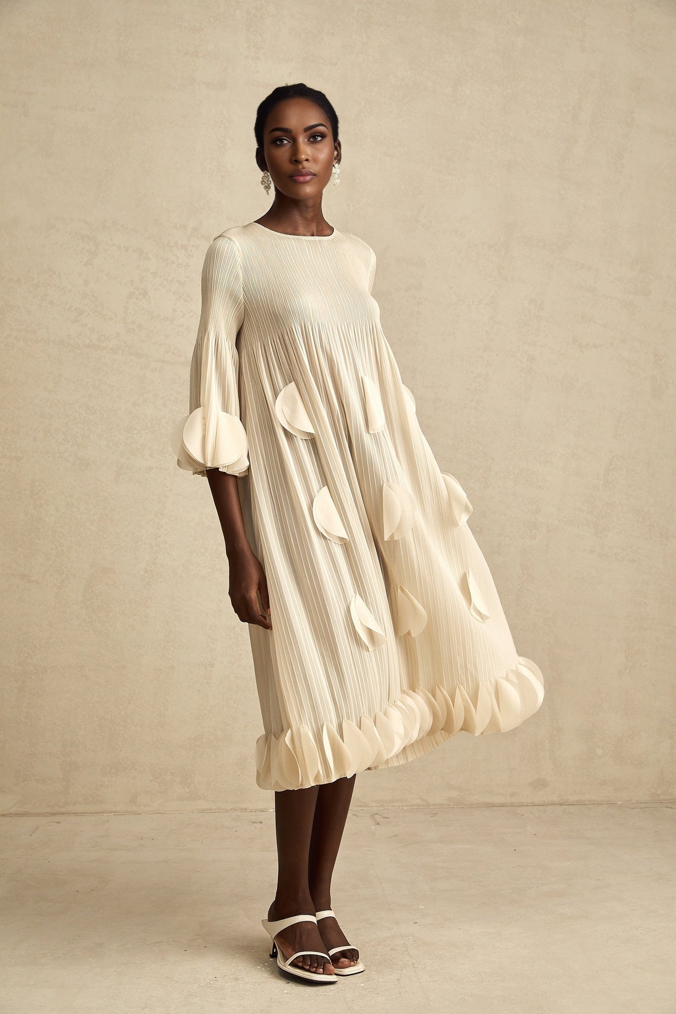 A woman wearing a light colored midi dress with intricate 3d floral petal pleats throughout the bodice and skirt standing in a neutral background