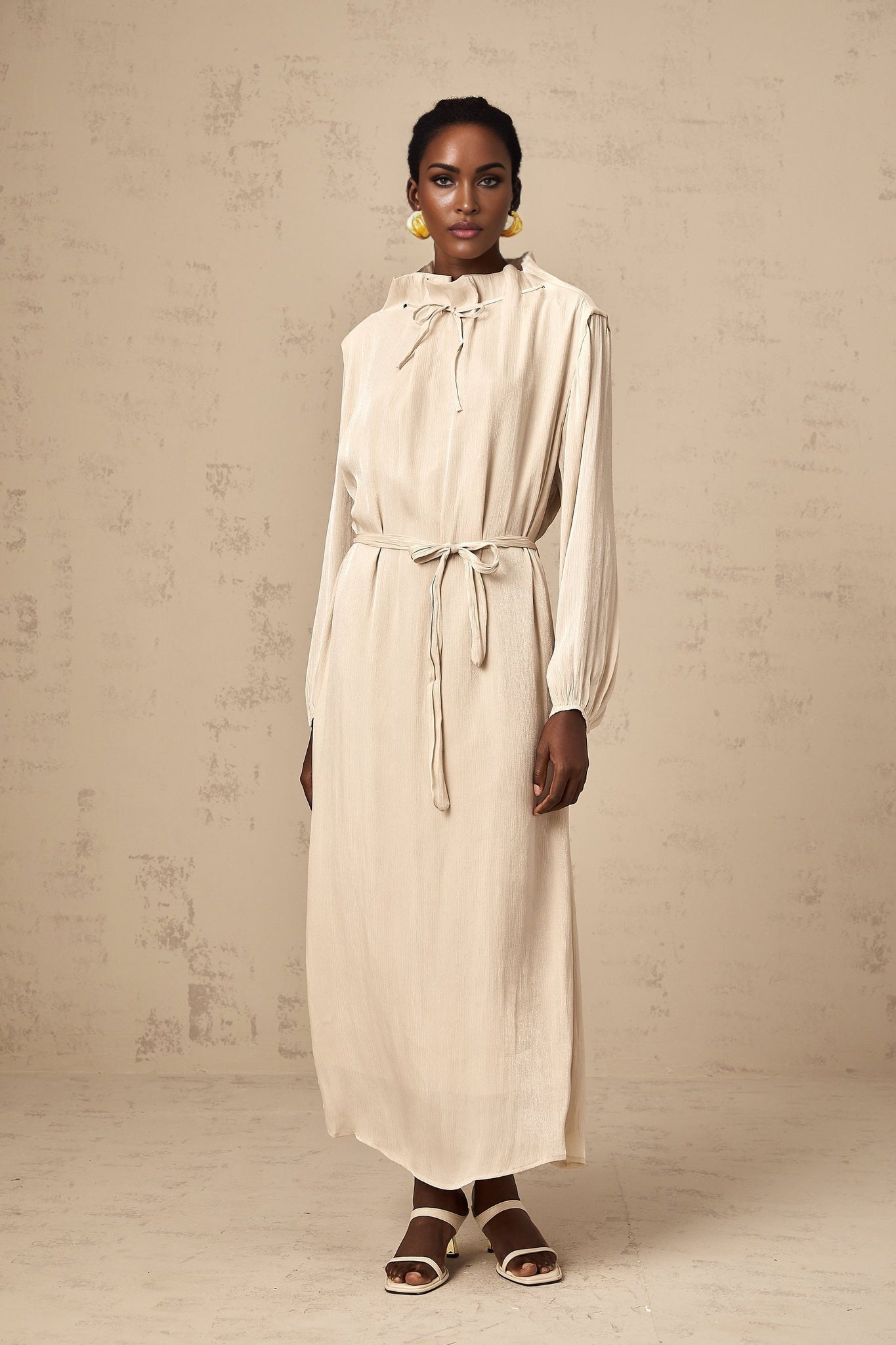 A woman wearing a light colored tulle midi dress with a self tie waist standing against a plain background