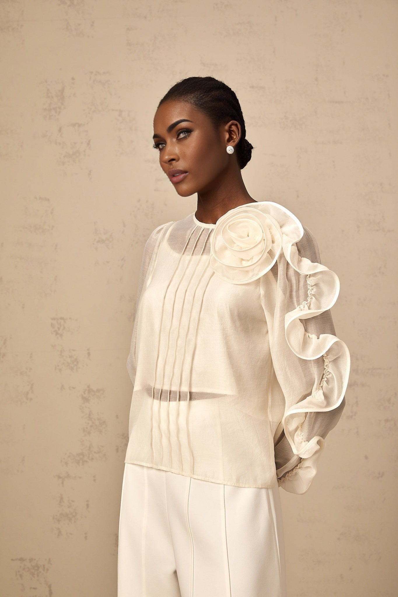A woman wearing a lightcolored sheer blouse with delicate three dimensional floral details standing against a neutral background