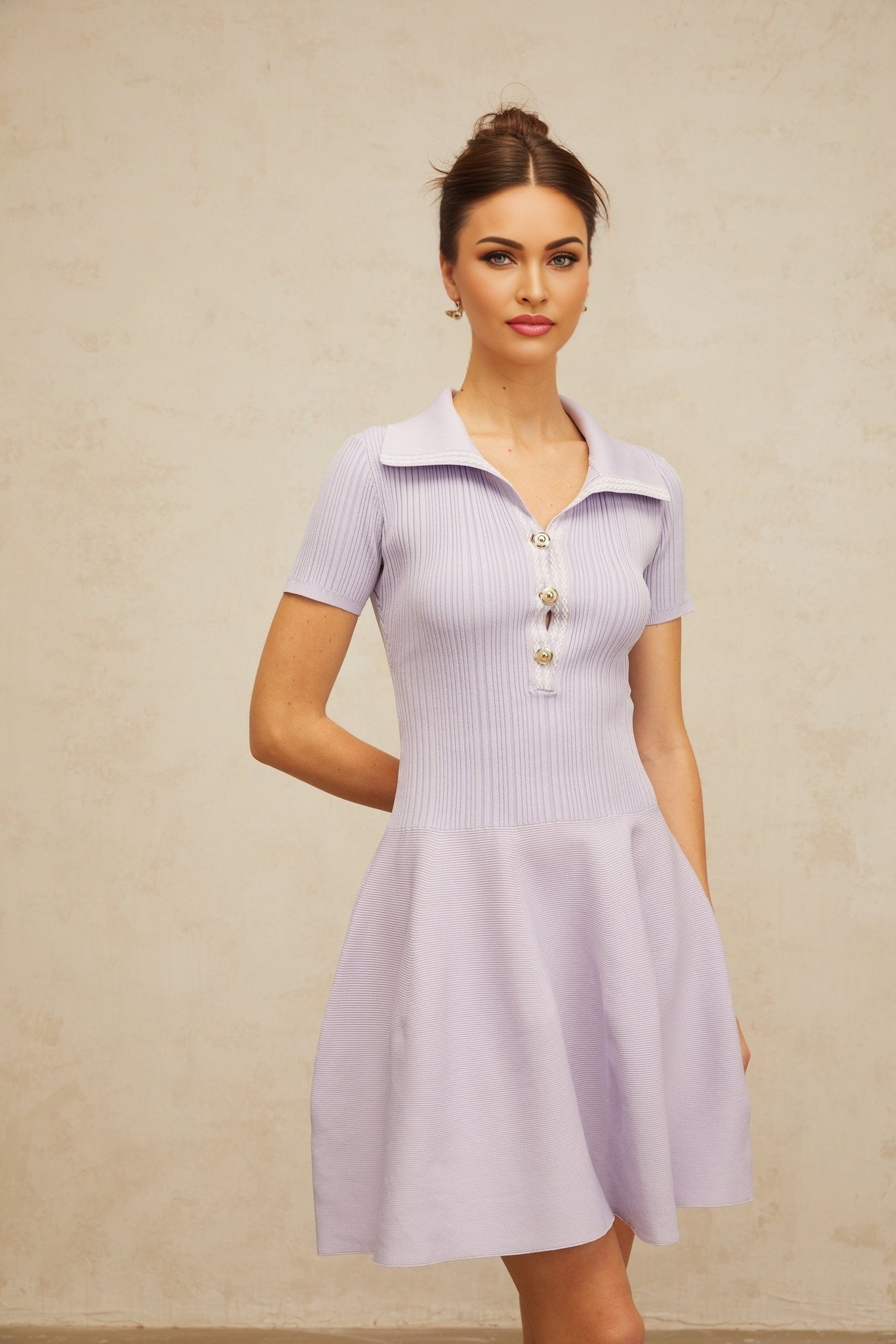 A woman wearing a lilac colored mini dress with a flared skirt and shirred bodice made of knit fabric the dress has short sleeves and falls above the knee